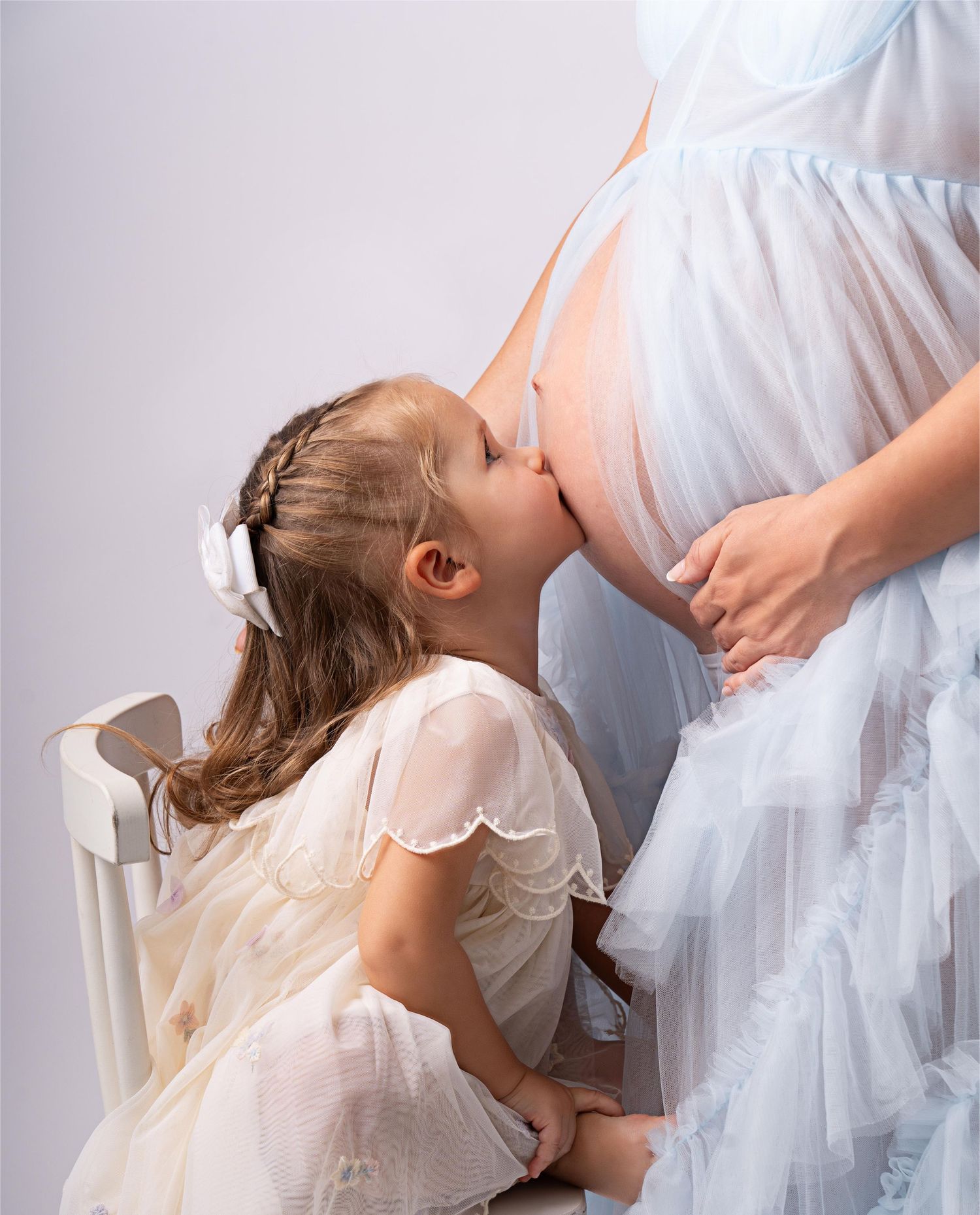 A young child in a white dress kisses a pregnant belly while kneeling on a chair in a tender family moment.