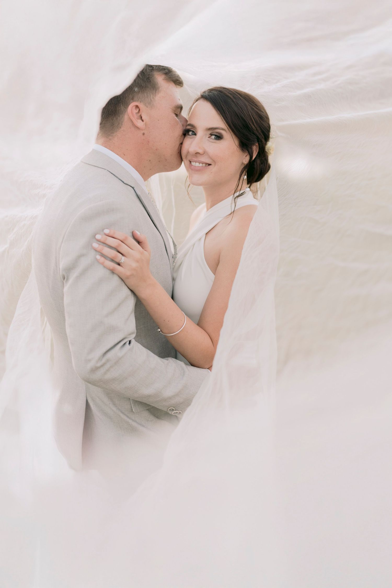 Smiling bride in white dress embraced by groom in grey suit under billowing wedding veil.