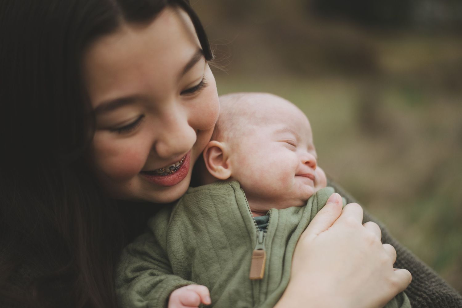A sleeping newborn baby in a green jacket is held lovingly against a girl wearing a dark sweater outdoors.