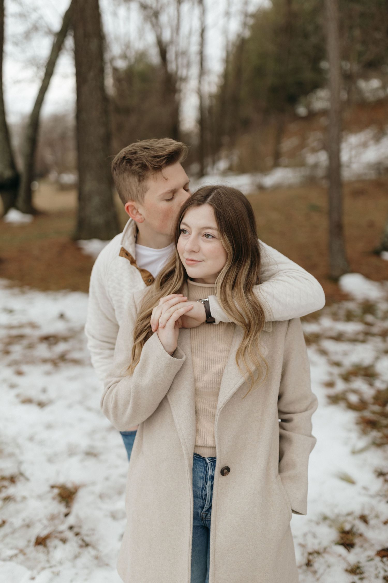 Cozy winter engagement photo with couple in neutral sweaters against snowy landscape.