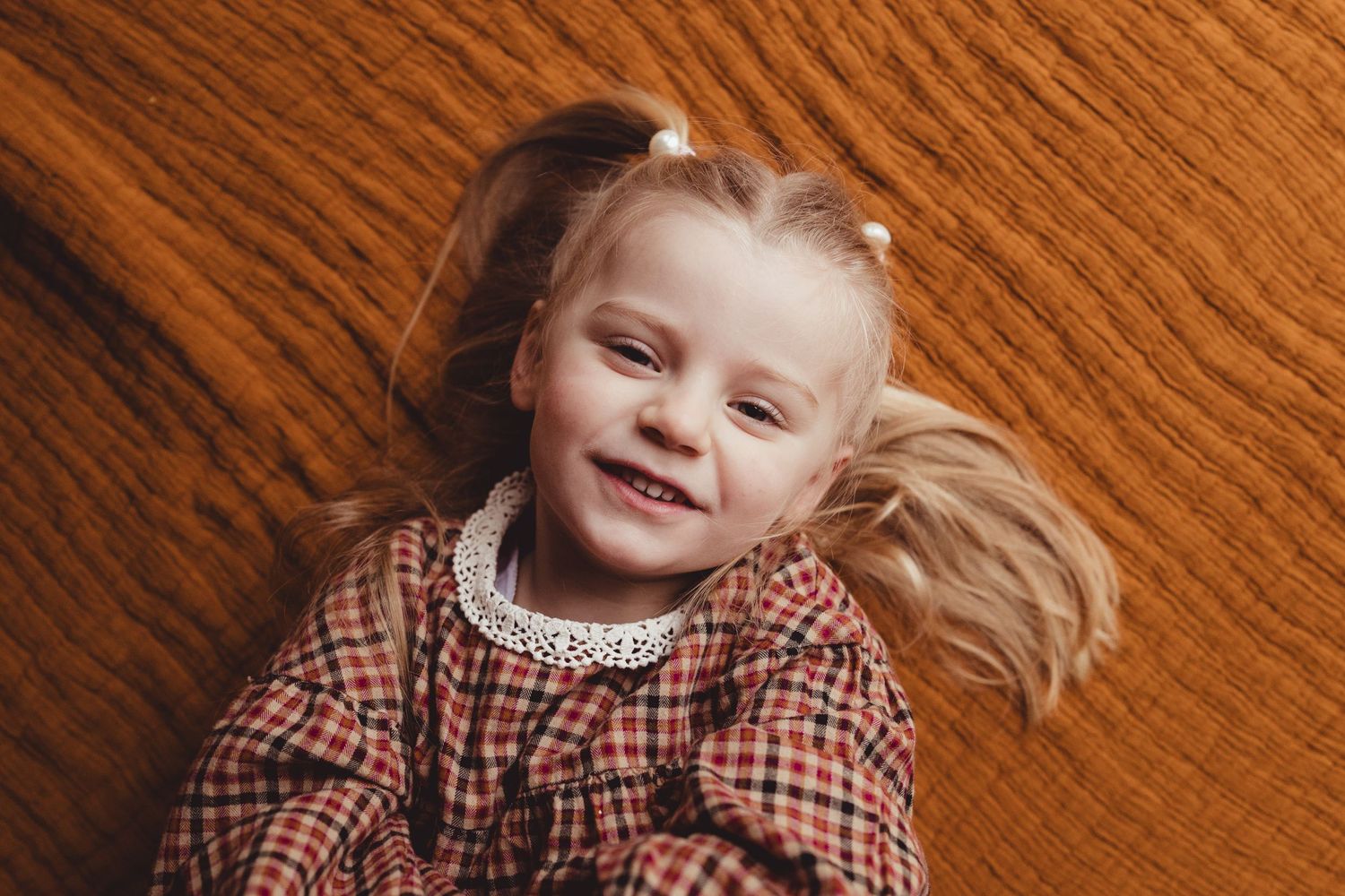 A child with blonde hair in a plaid dress smiles while lying on a wood grain background.