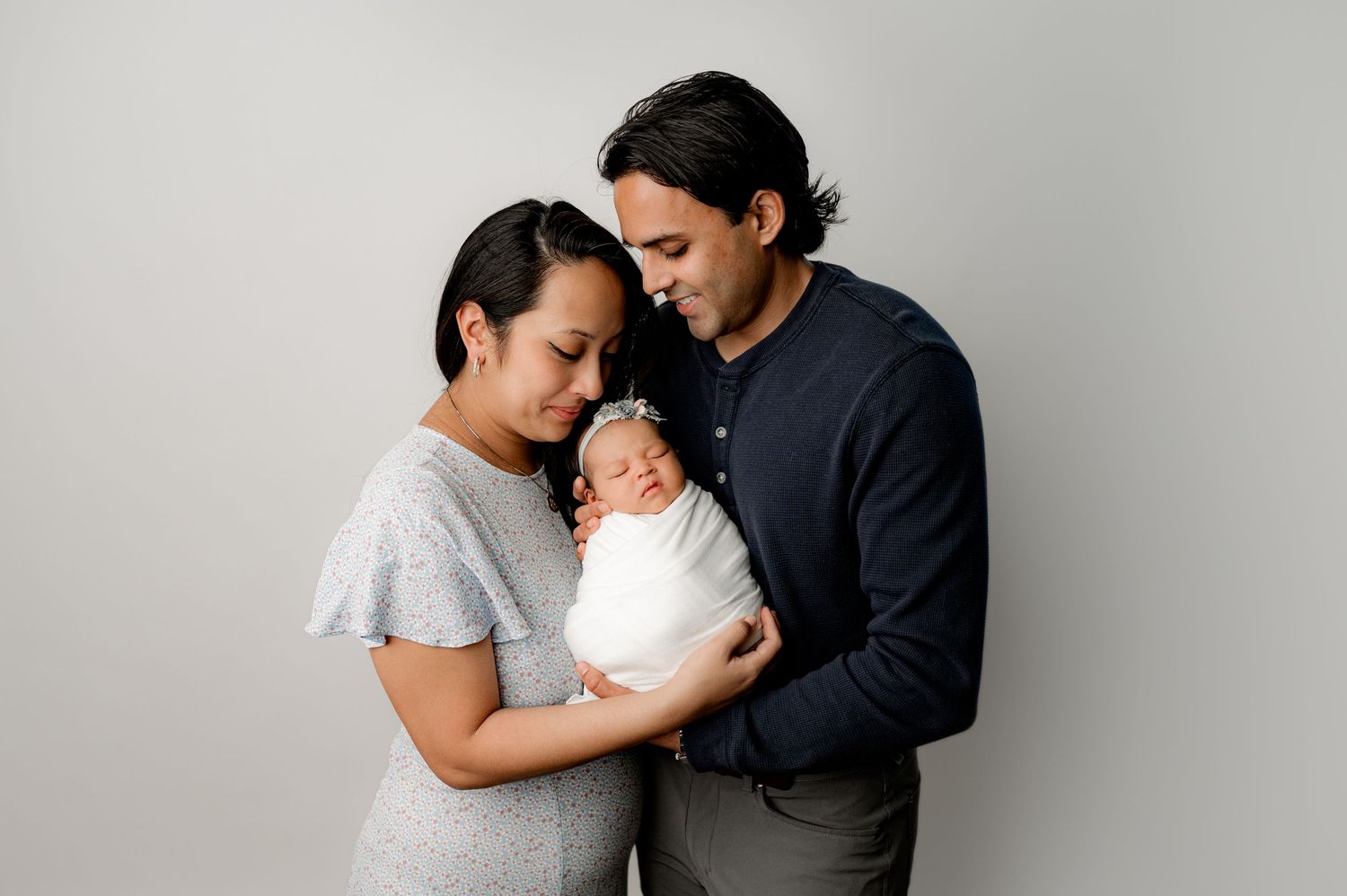 Family portrait with parents tenderly holding newborn against grey backdrop.