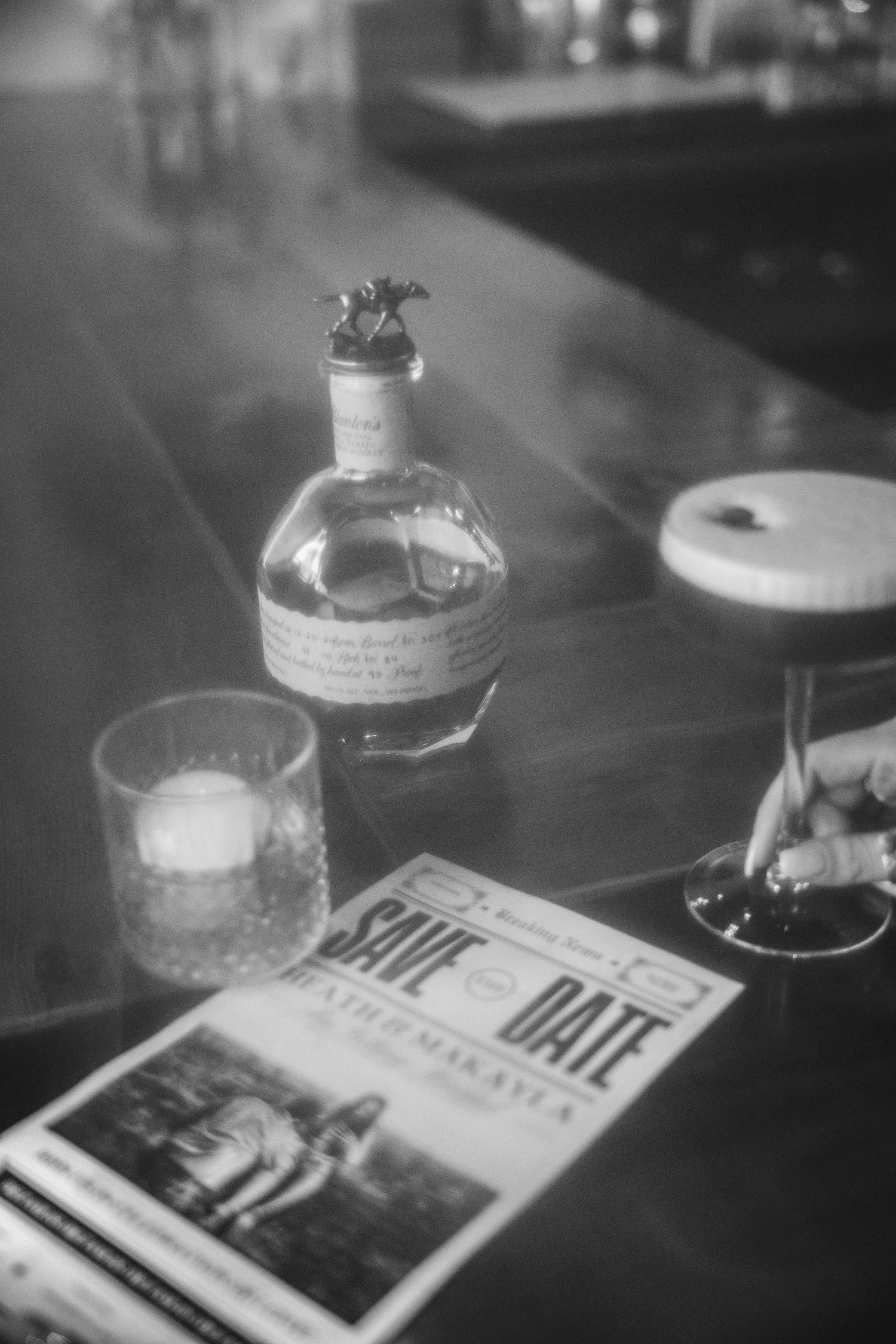 Black and white photo of a table setting with a decorative bottle, drink glass, and printed materials in moody lighting.