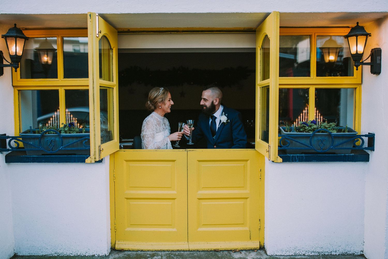 A couple shares an intimate moment at a bright yellow dutch door with lanterns and flower boxes during their wedding day.