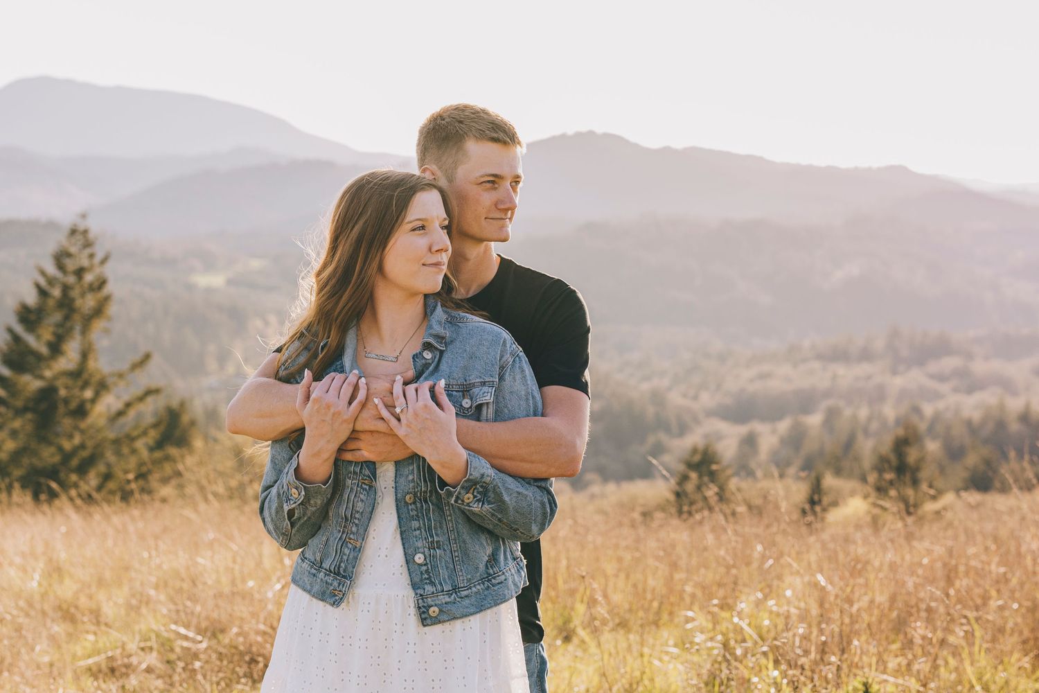 A couple embraces while standing in a golden meadow with mountain views in the background.