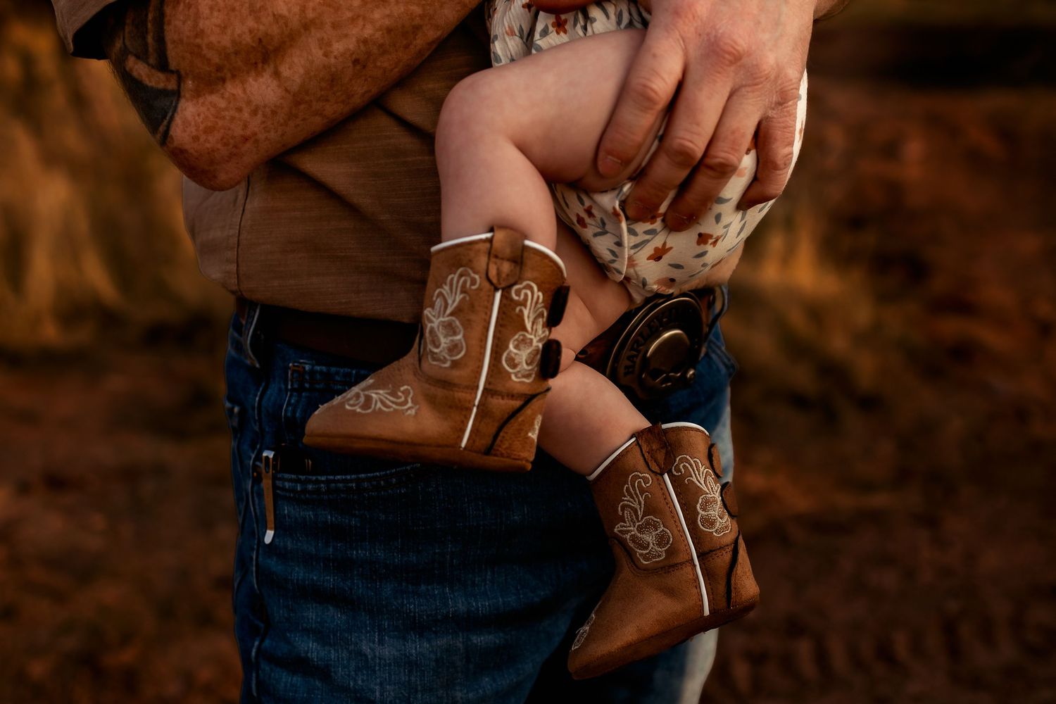 Close up of tiny brown leather cowboy boots with embroidered designs being held by someone wearing blue jeans.