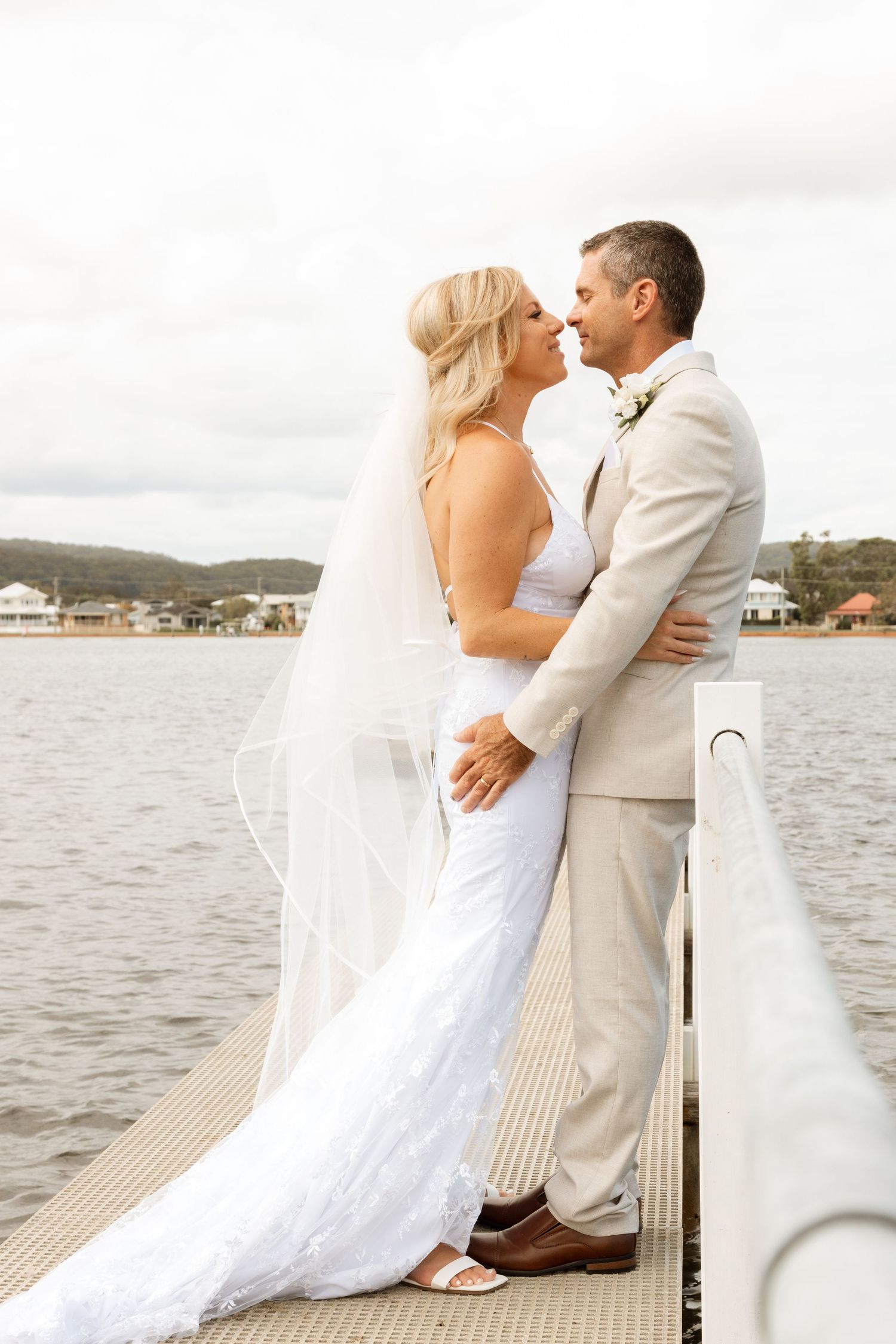 A couple shares an intimate moment on a dock by the water, with the bride in white and groom in beige.