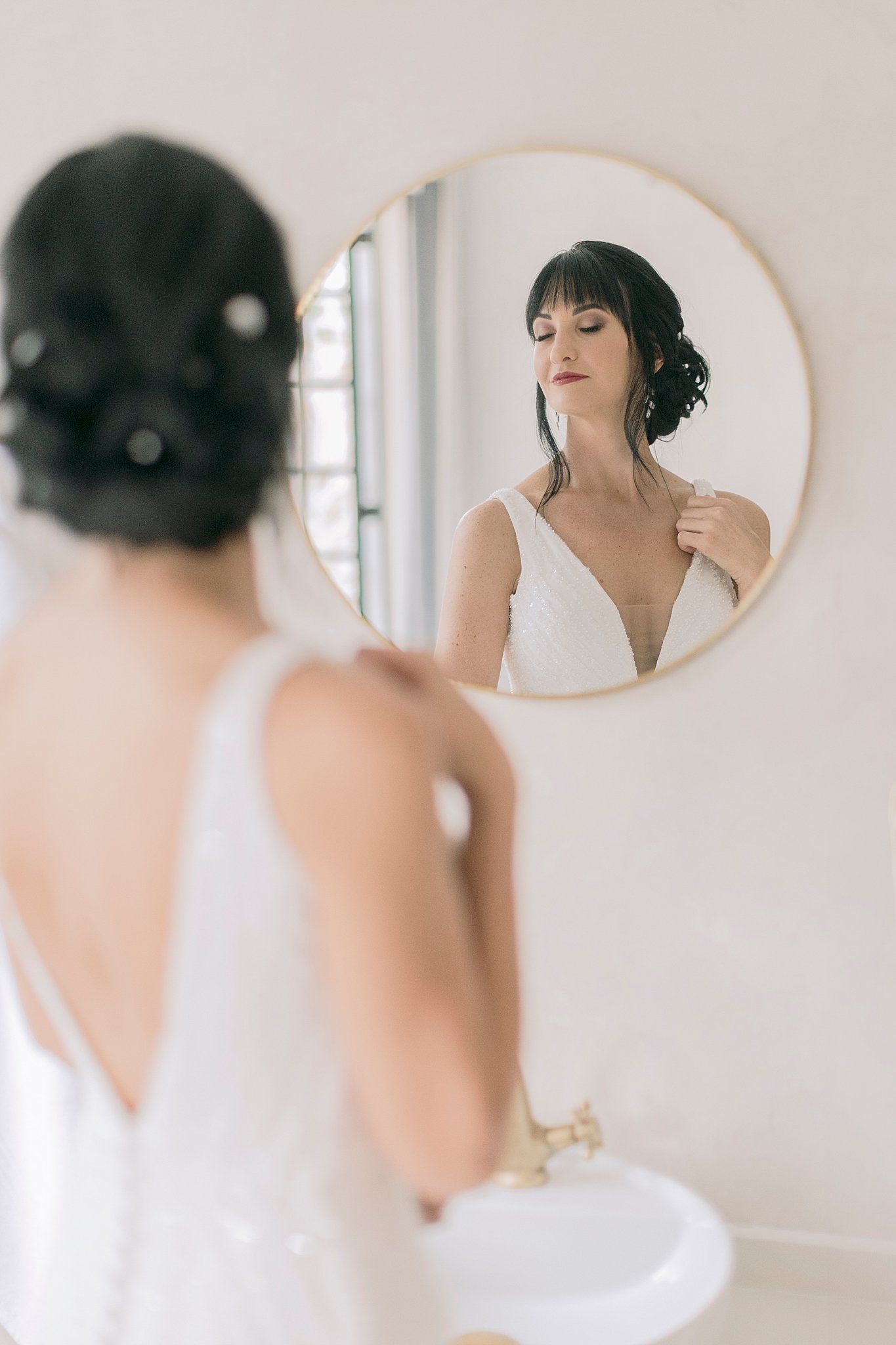 Beautiful dark-haired bride in white gown admiring herself in round gold mirror on wedding day.