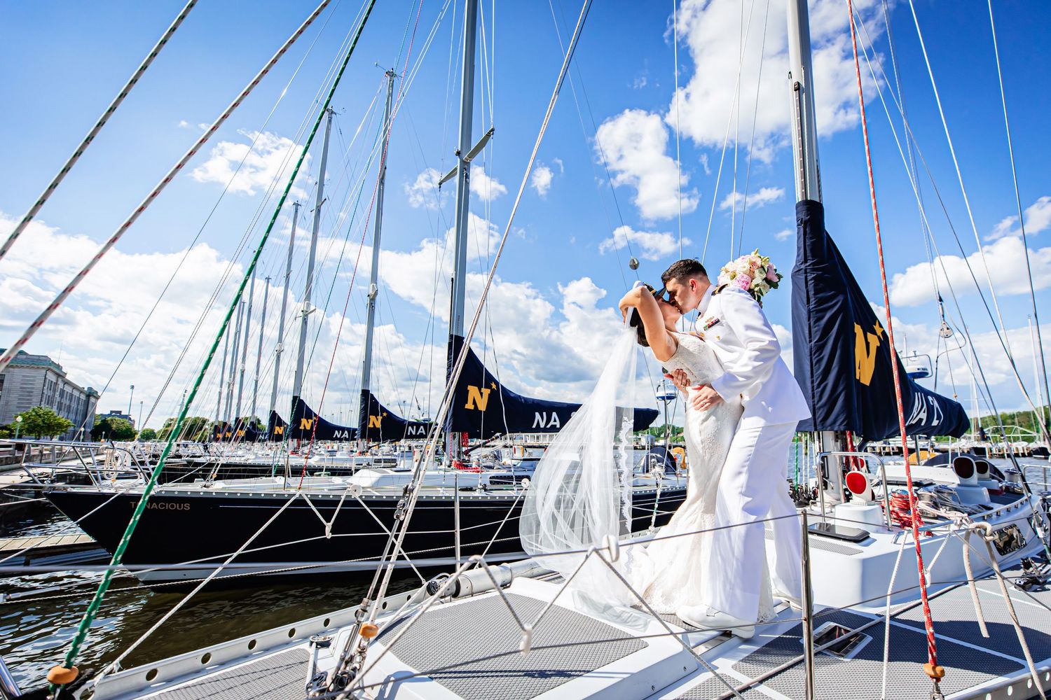 A couple in wedding attire embraces on the deck of a sailboat at a marina on a sunny summer day.