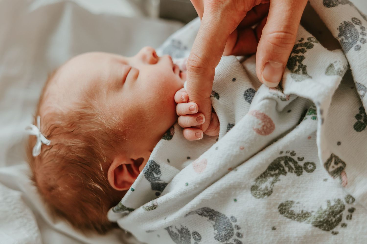 A baby wearing a gray star-print outfit plays on a white quilted surface while an adult's hands reach into frame.