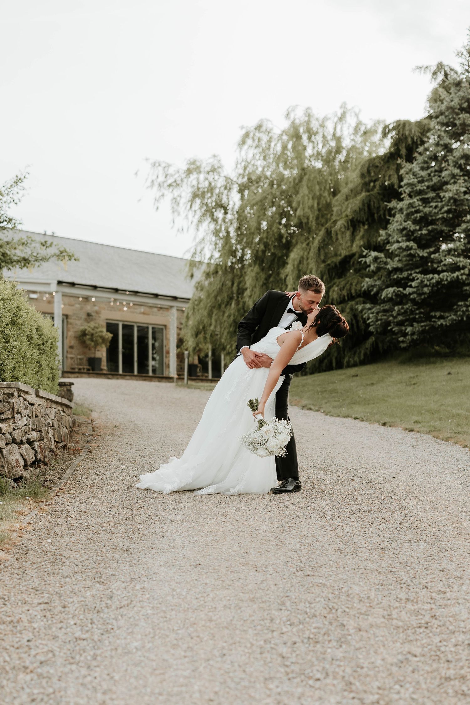 A couple in wedding attire dances together on a gravel path surrounded by lush greenery on their special day.