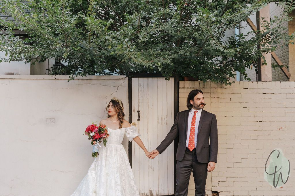 A couple stands against a brick wall covered in lush green ivy, with the bride wearing an elegant lace wedding gown.