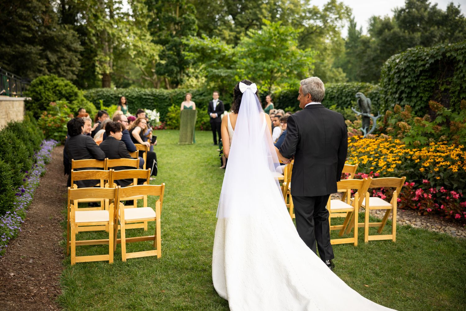 A beautiful outdoor garden wedding ceremony with wooden chairs and colorful flower beds lining the aisle. Pittsburgh National Aviary