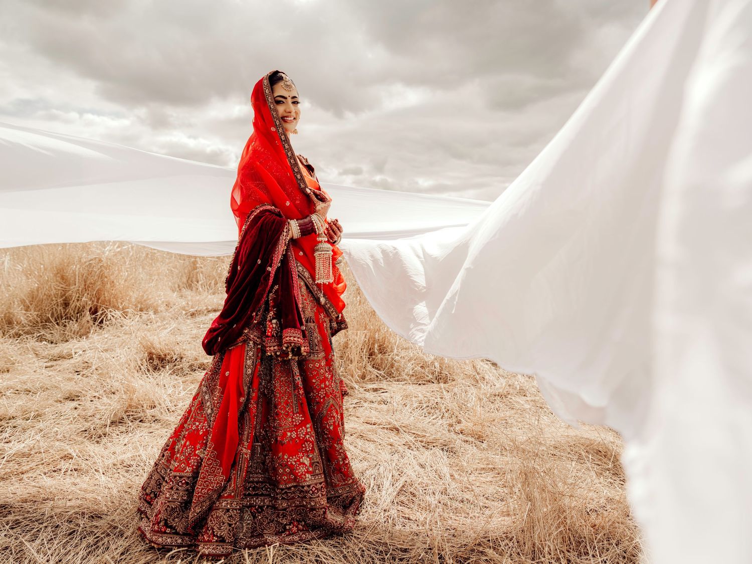 A bride in a vibrant red traditional Indian lehenga poses in a field while her veil flows dramatically in the wind.