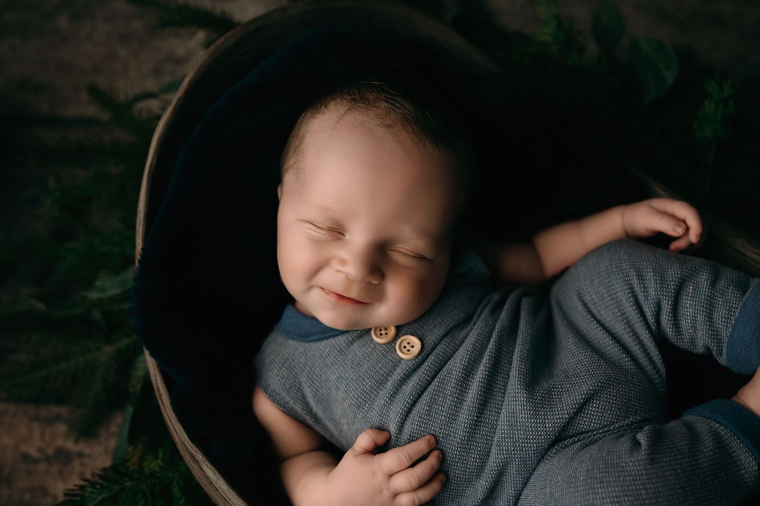 A joyful baby wearing gray clothing lies back with a big smile on a dark background.