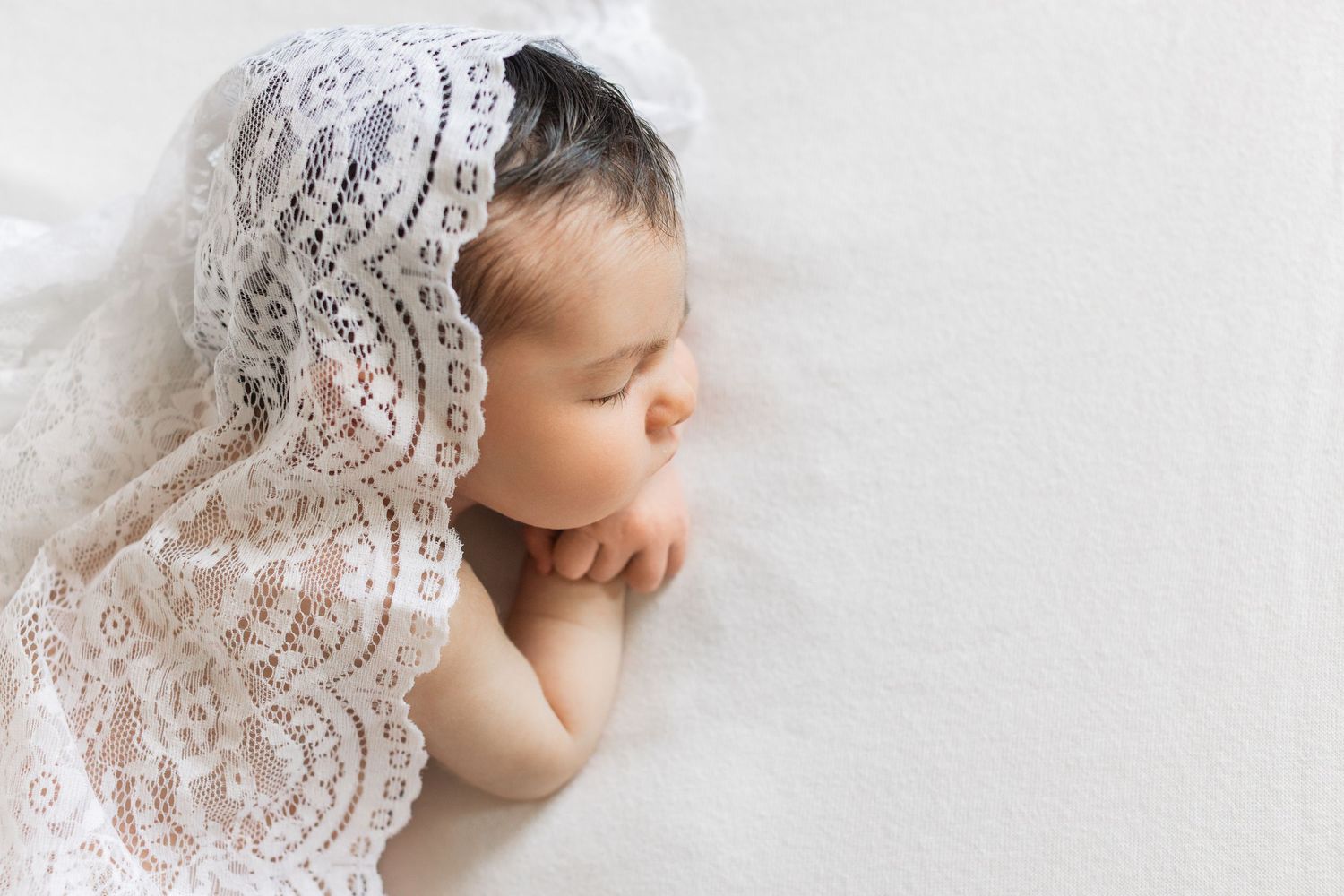 A delicate white lace overlay drapes over a sleeping newborn on a soft white background during a photography session.