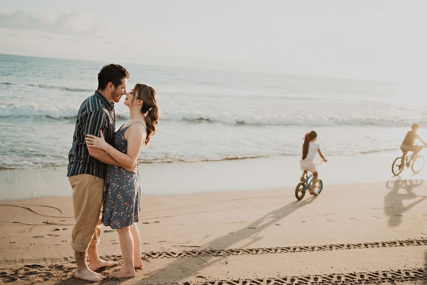 Couple sharing a kiss on the beach, with shadows of children on bicycles playfully pointing during a pre-wedding shoot
