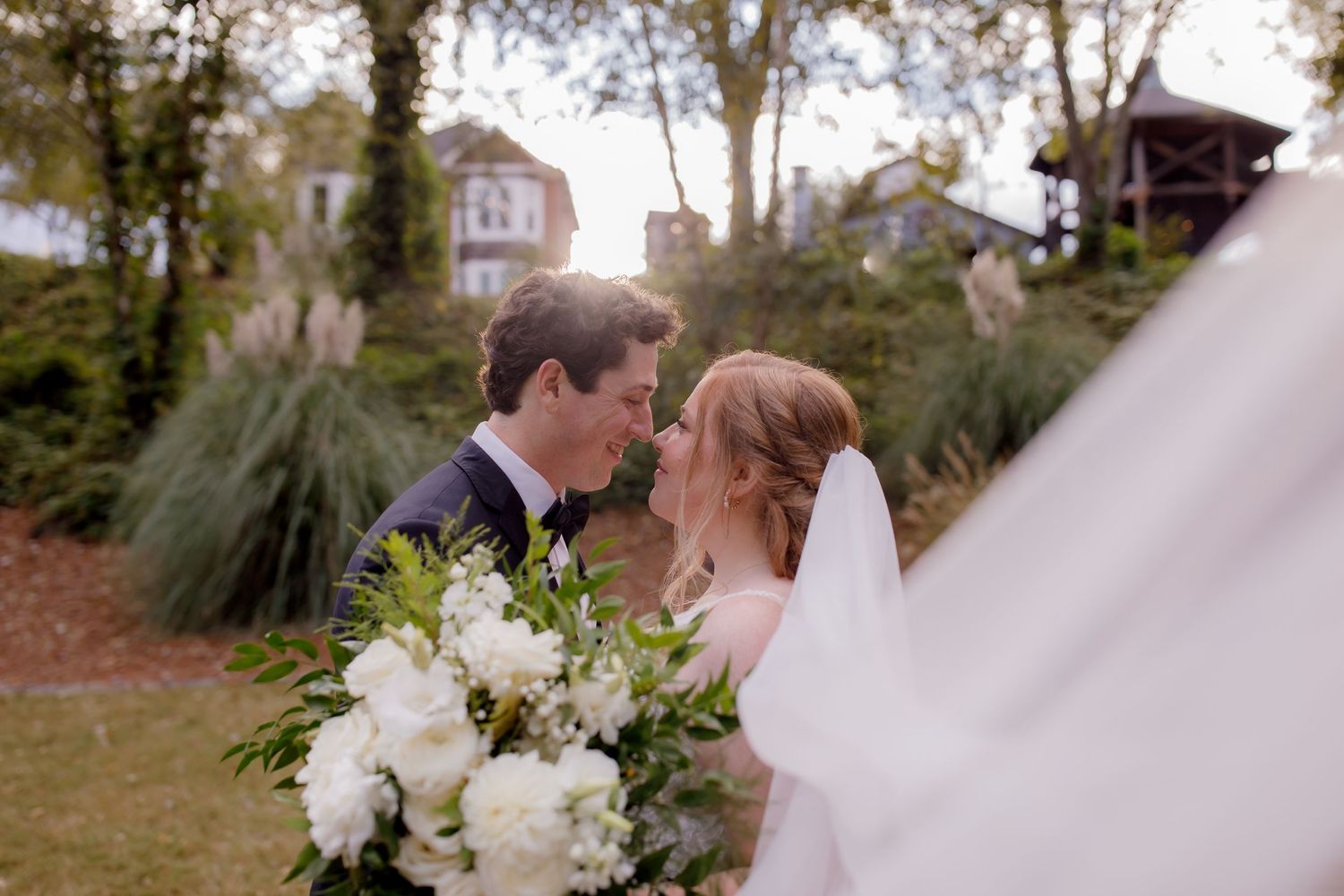 A couple shares a romantic moment outdoors with a white floral bouquet against a sunlit garden backdrop.