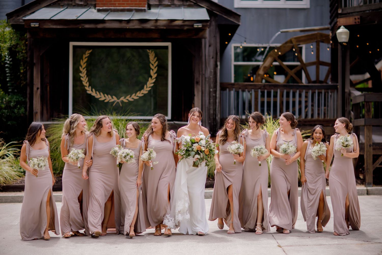 Wedding party in matching blush gowns posing outdoors with rustic wooden backdrop.
