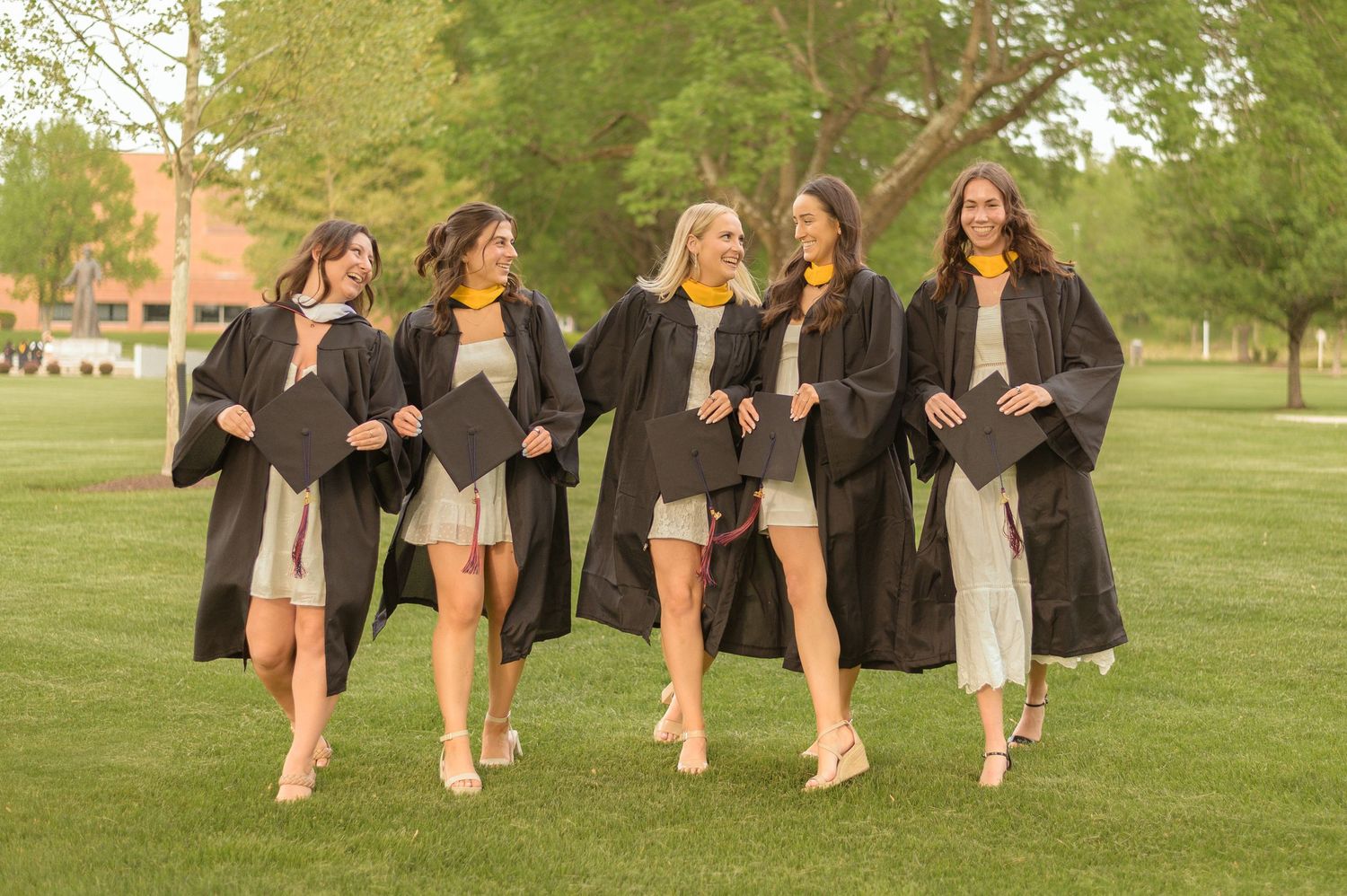 Group in light-colored dresses standing on grass against beige wall tossing graduation caps in celebration.