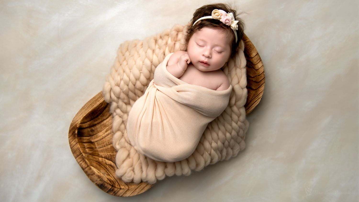 Sleeping newborn baby swaddled in beige fabric lies peacefully in a woven basket on neutral backdrop.