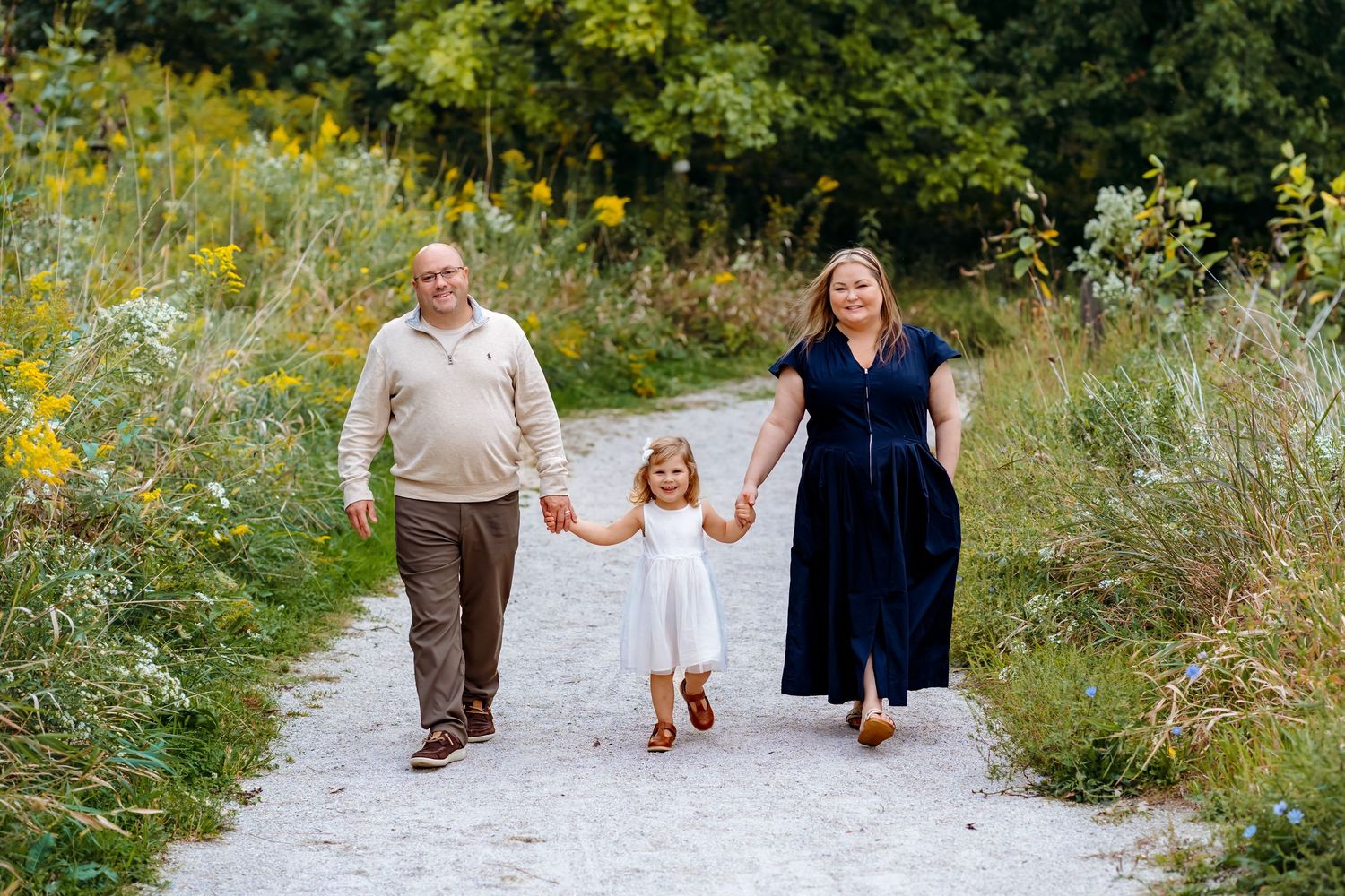 A family walking together on a gravel path surrounded by wildflowers and tall grasses on a sunny day.