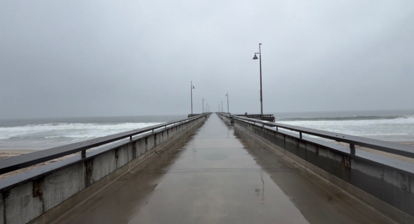 A long wooden pier extends into the foggy ocean on a cloudy, overcast day at the beach.