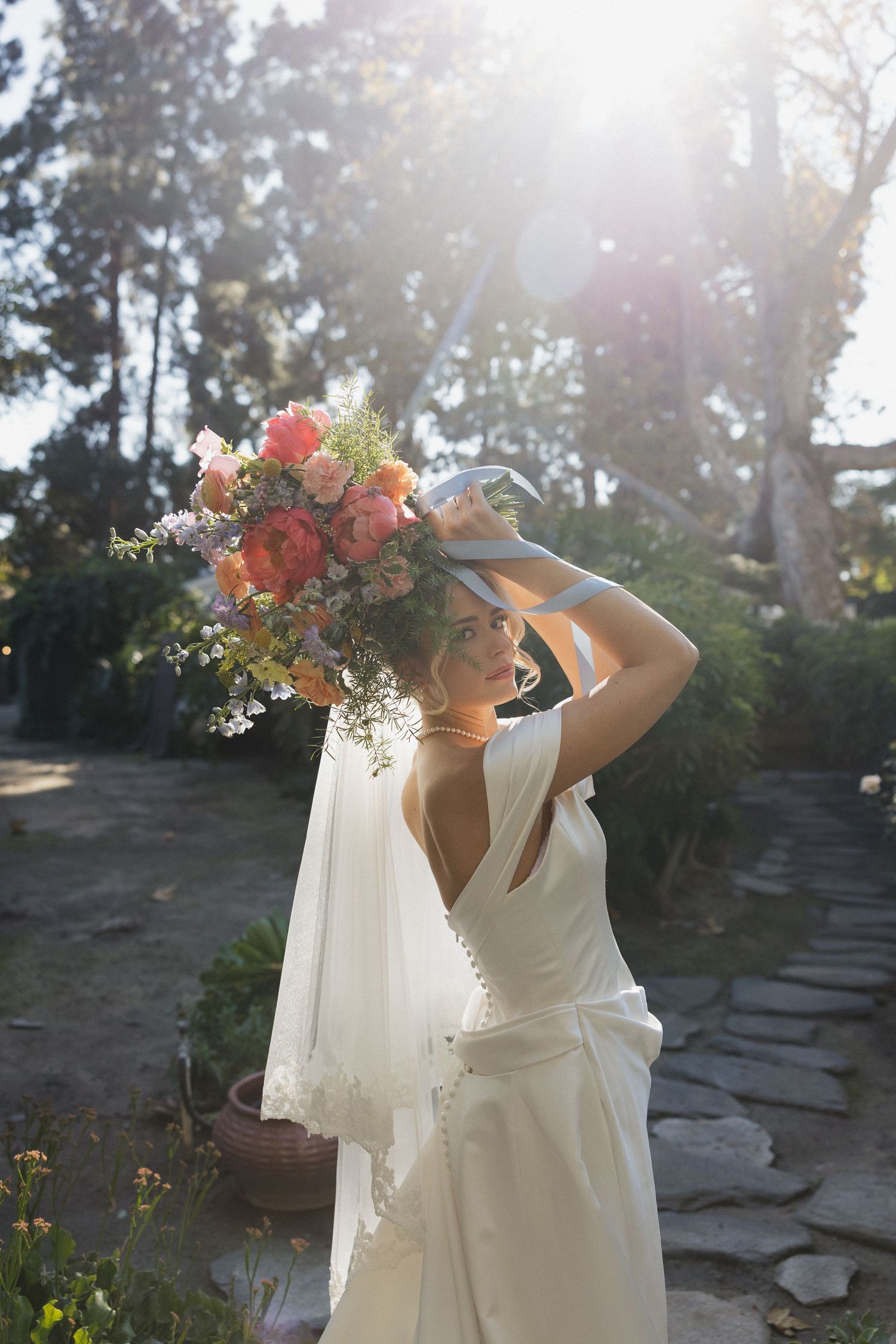 A white flowy dress catches sunlight in a garden setting with stone pathways and delicate flower bouquets.