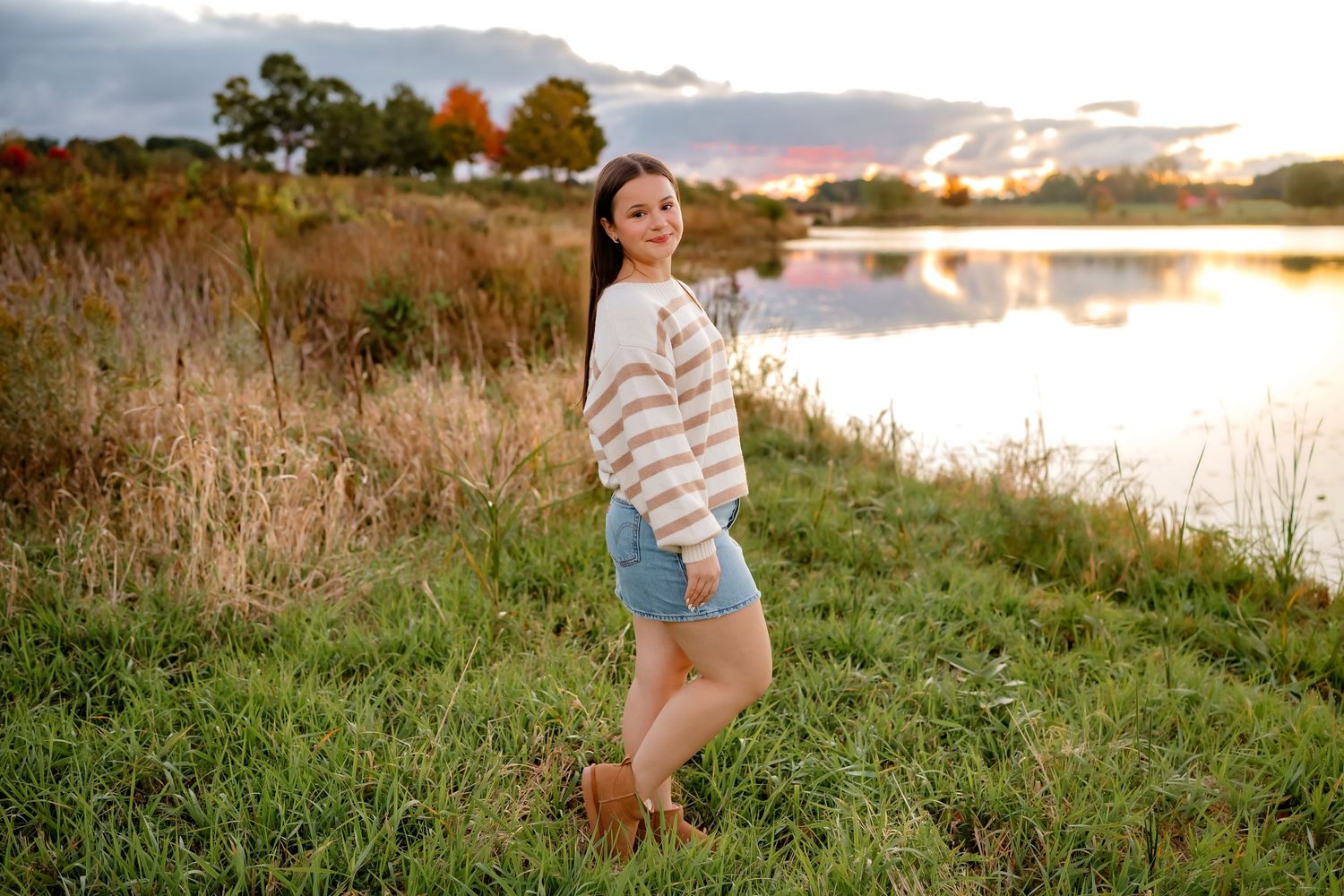A person sits in tall grass near a tranquil lake during golden hour sunset, creating a peaceful outdoor scene.