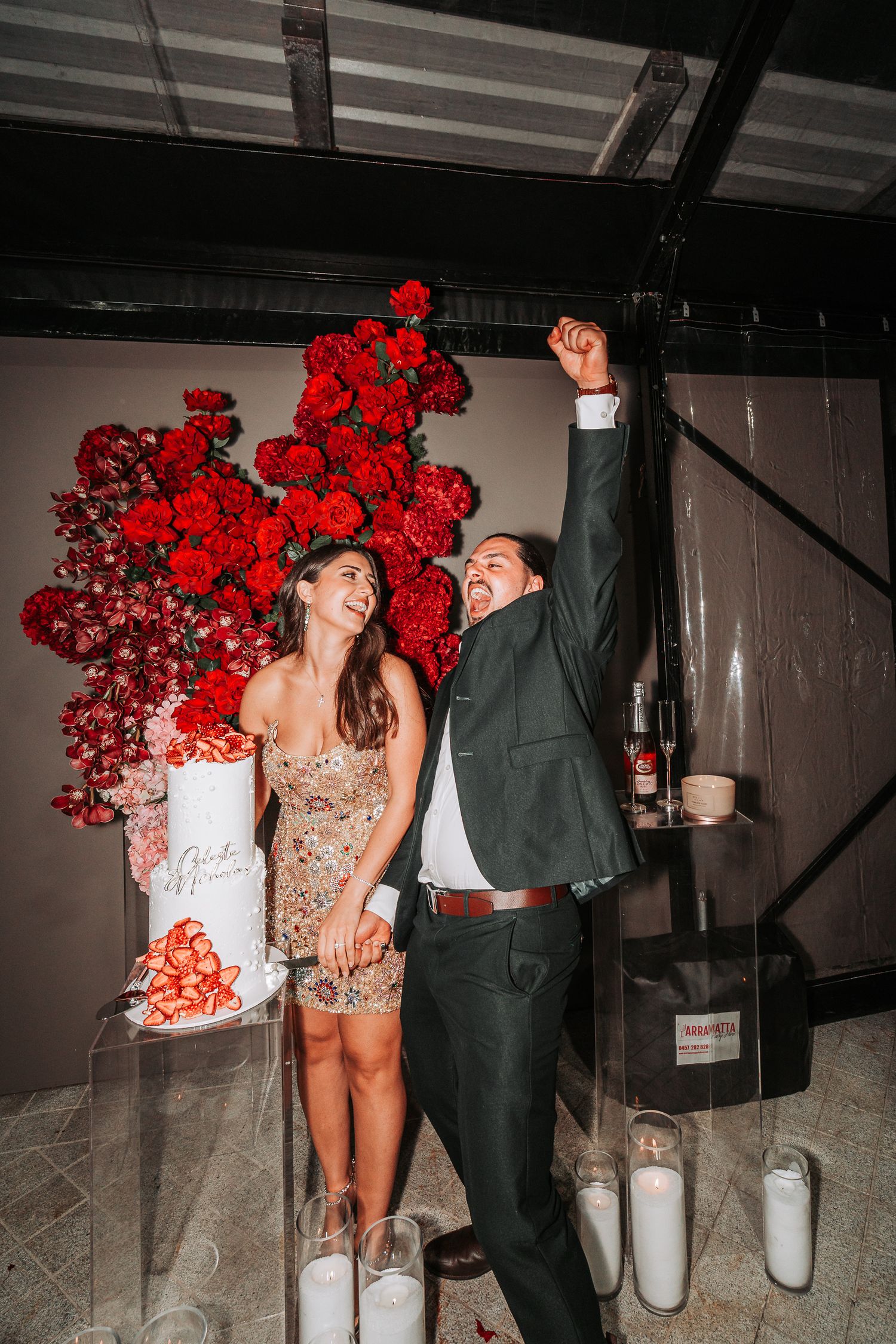 A couple poses with vibrant red roses in a dimly lit industrial space wearing formal attire and sharing playful moments.