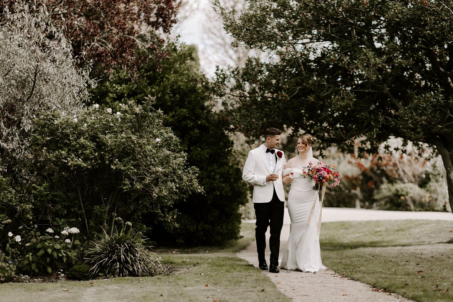 A couple walks hand in hand through a lush garden pathway during an outdoor wedding celebration.