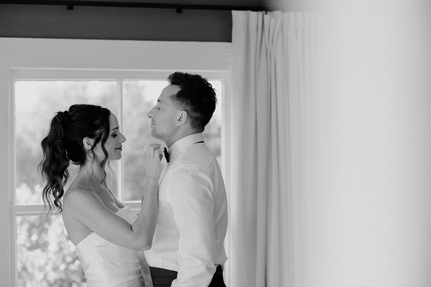 Black and white image of a bride helping the groom with his bowtie and sharing an intimate moment beside a window by August & Vine
