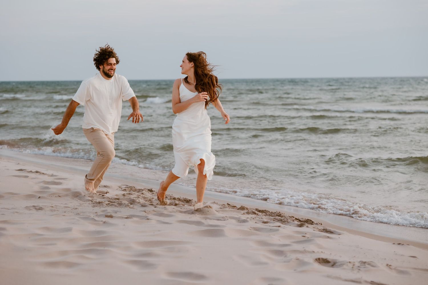 Two people wearing white outfits walking and splashing together along the shoreline at sunset.