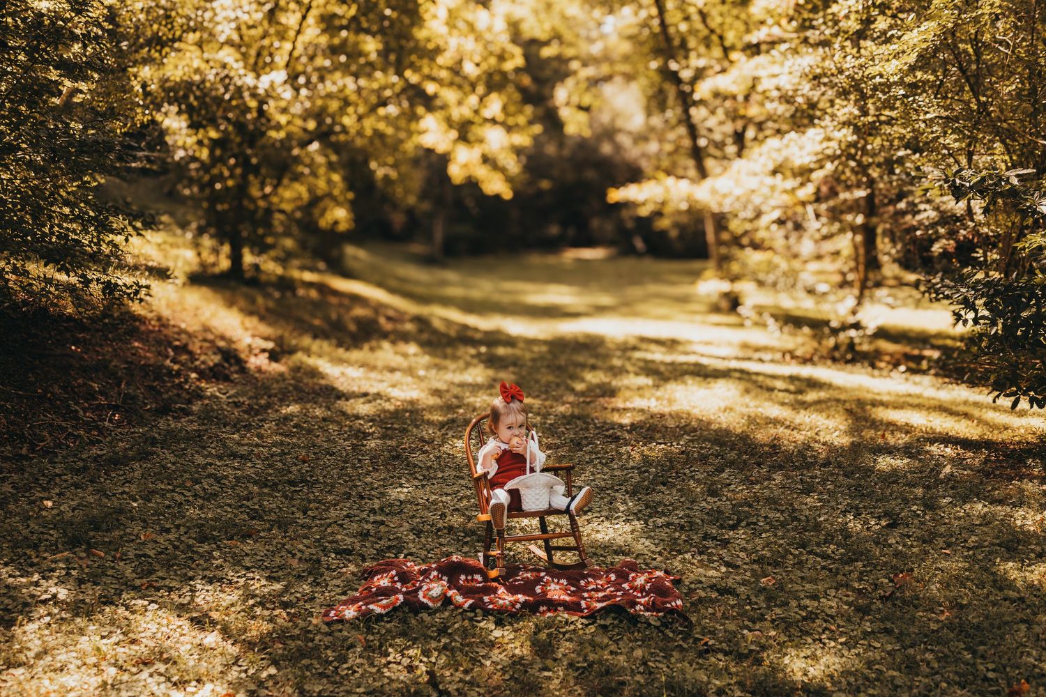 Toddler in red dress sitting in rocking chair on colorful blanket surrounded by autumn leaves in forest.