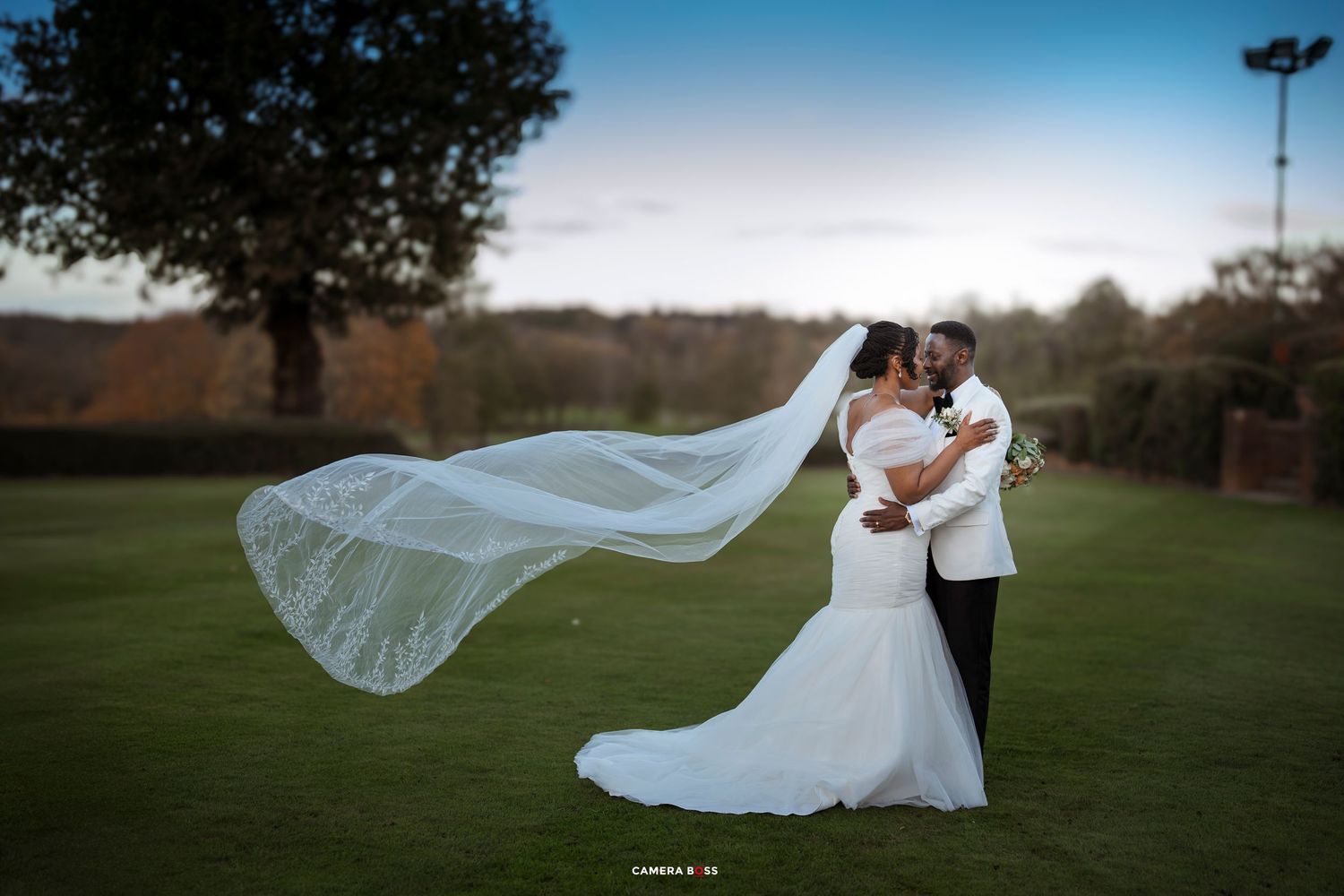 A romantic wedding couple embraces on a grassy field as the bride's veil flows dramatically in the wind at sunset.