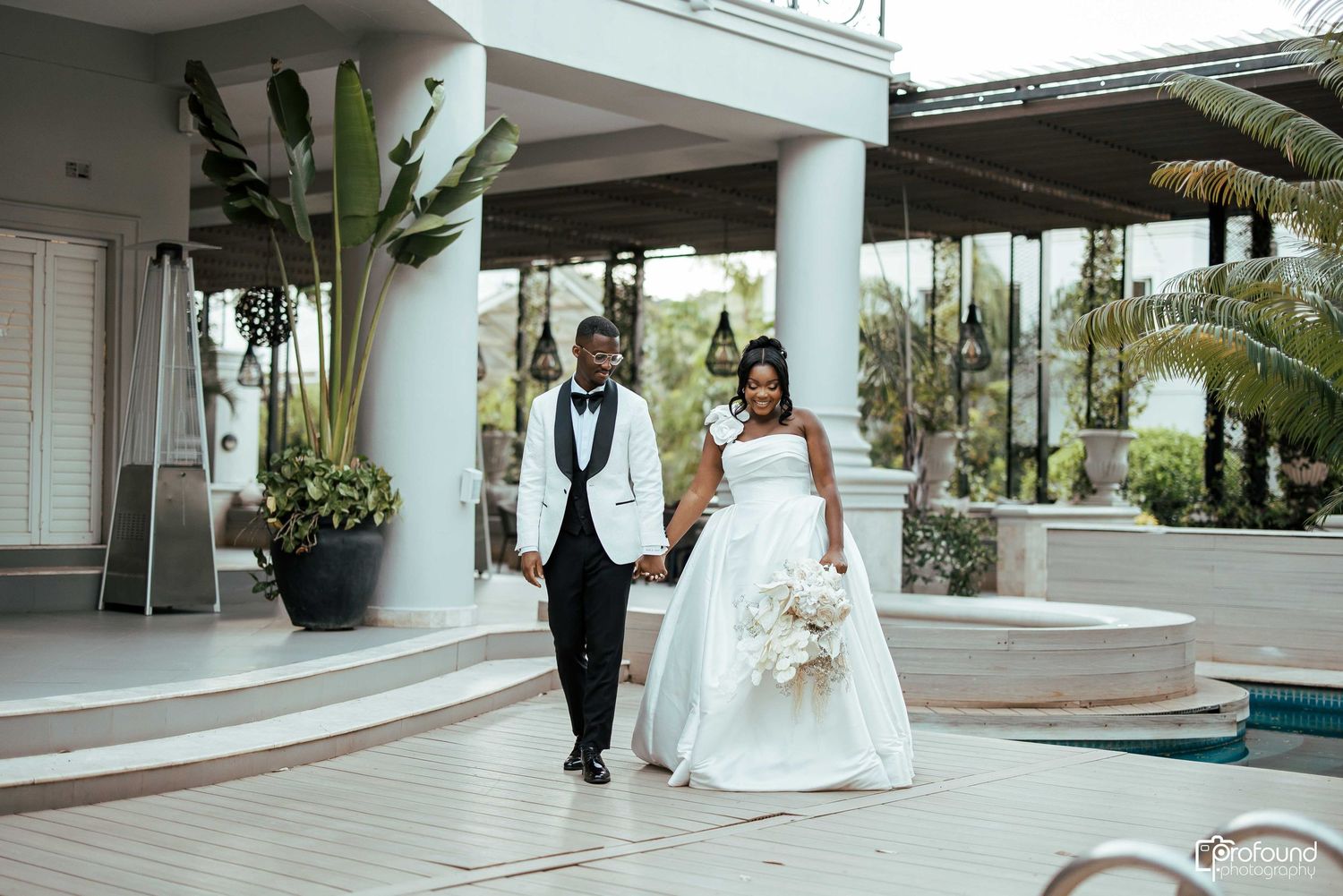 A bride and groom walk through an elegant hotel lobby with marble floors and white columns.