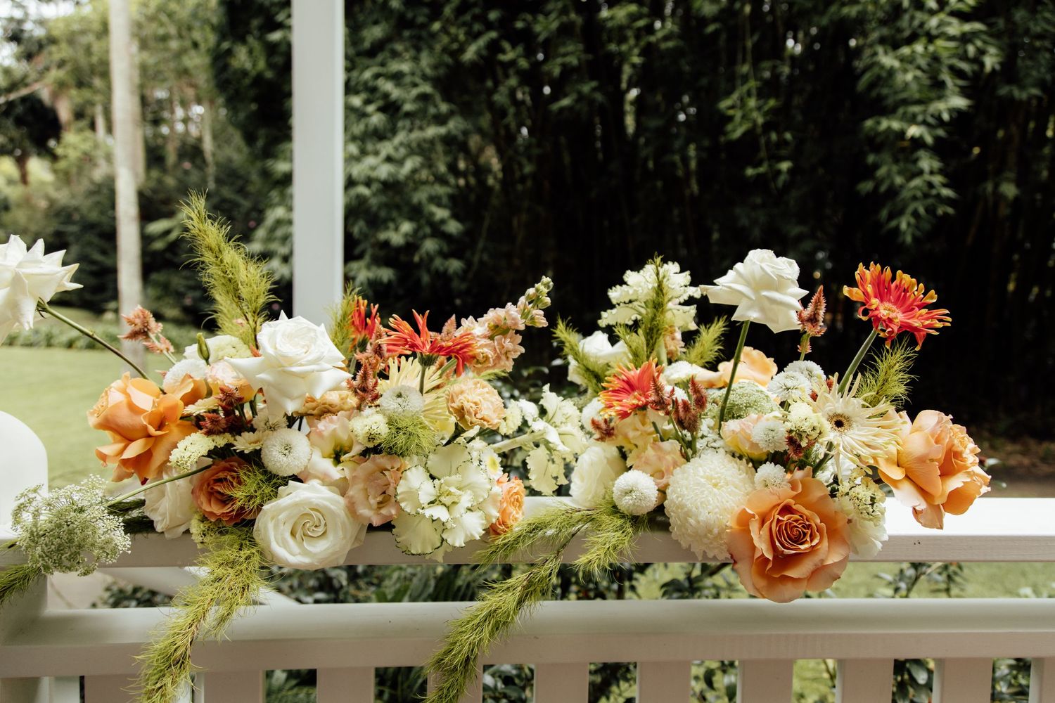 Floral arrangements with orange roses, white flowers, and greenery line a white railing in an outdoor garden setting.