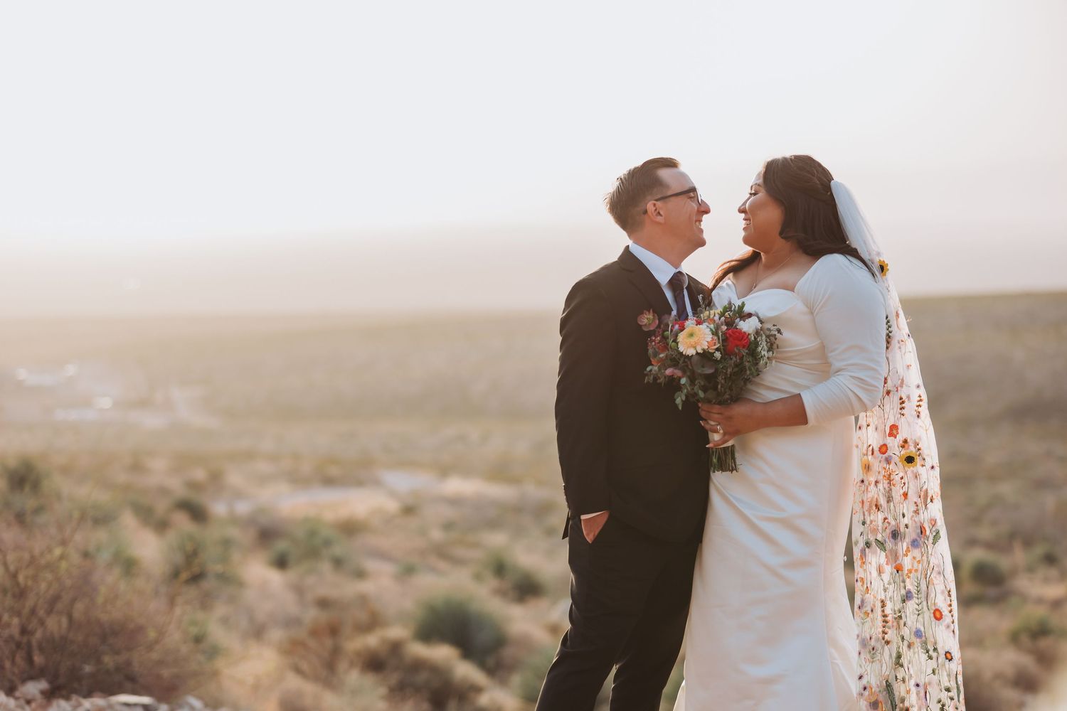 A couple in wedding attire embraces while standing on a hilltop overlooking a desert landscape at sunset.