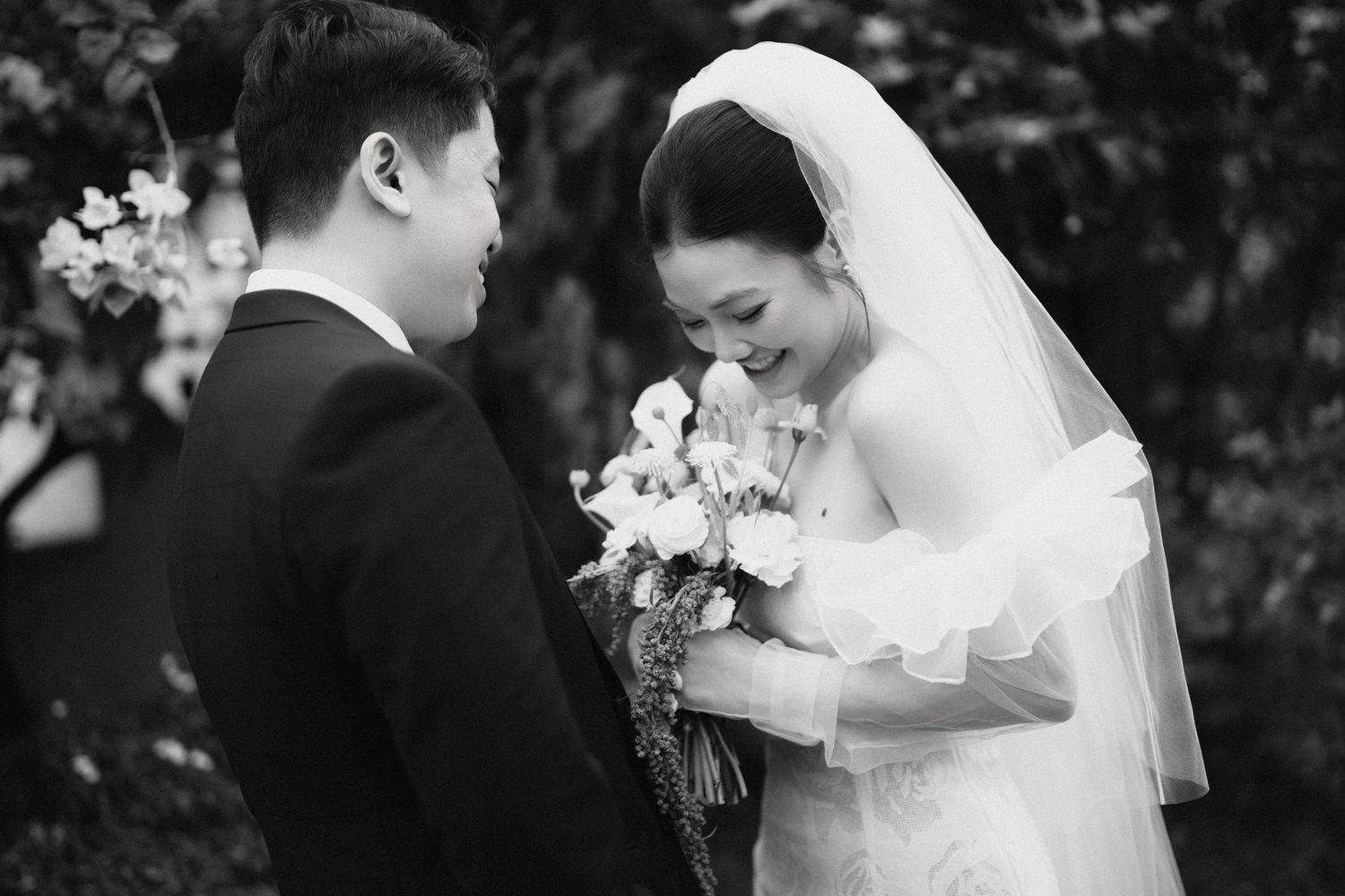 Black and white photo of Asian bride and groom sharing a tender moment, bride holding a floral bouquet outdoors.