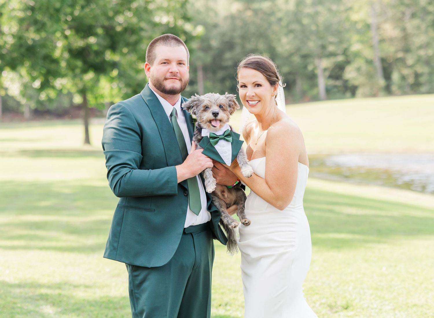 Newlywed couple poses with their small gray dog on a sunny golf course.
