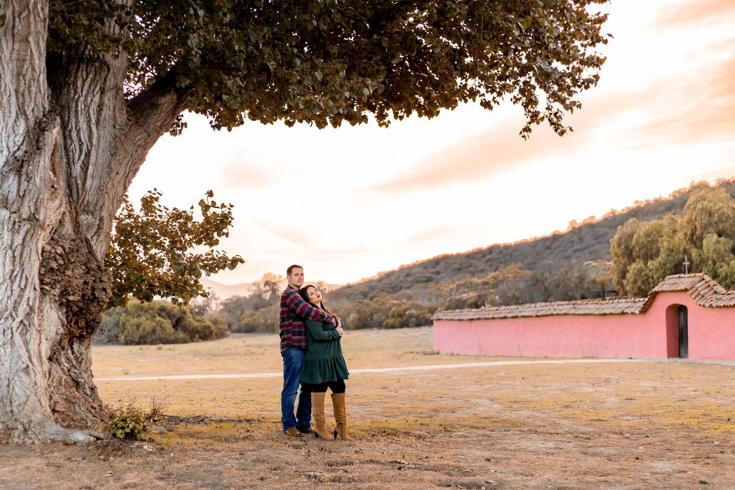 A couple stands together under a large tree in a serene autumn landscape with warm sunset lighting.