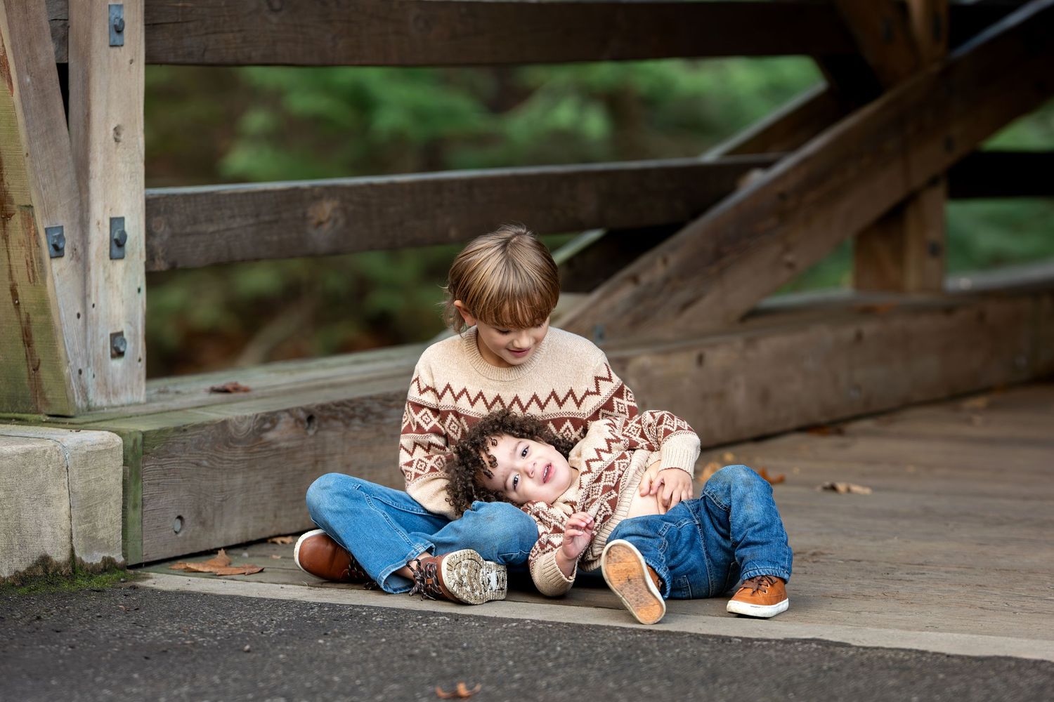 Two young children sit together on a wooden bridge deck, one resting on the other's lap.