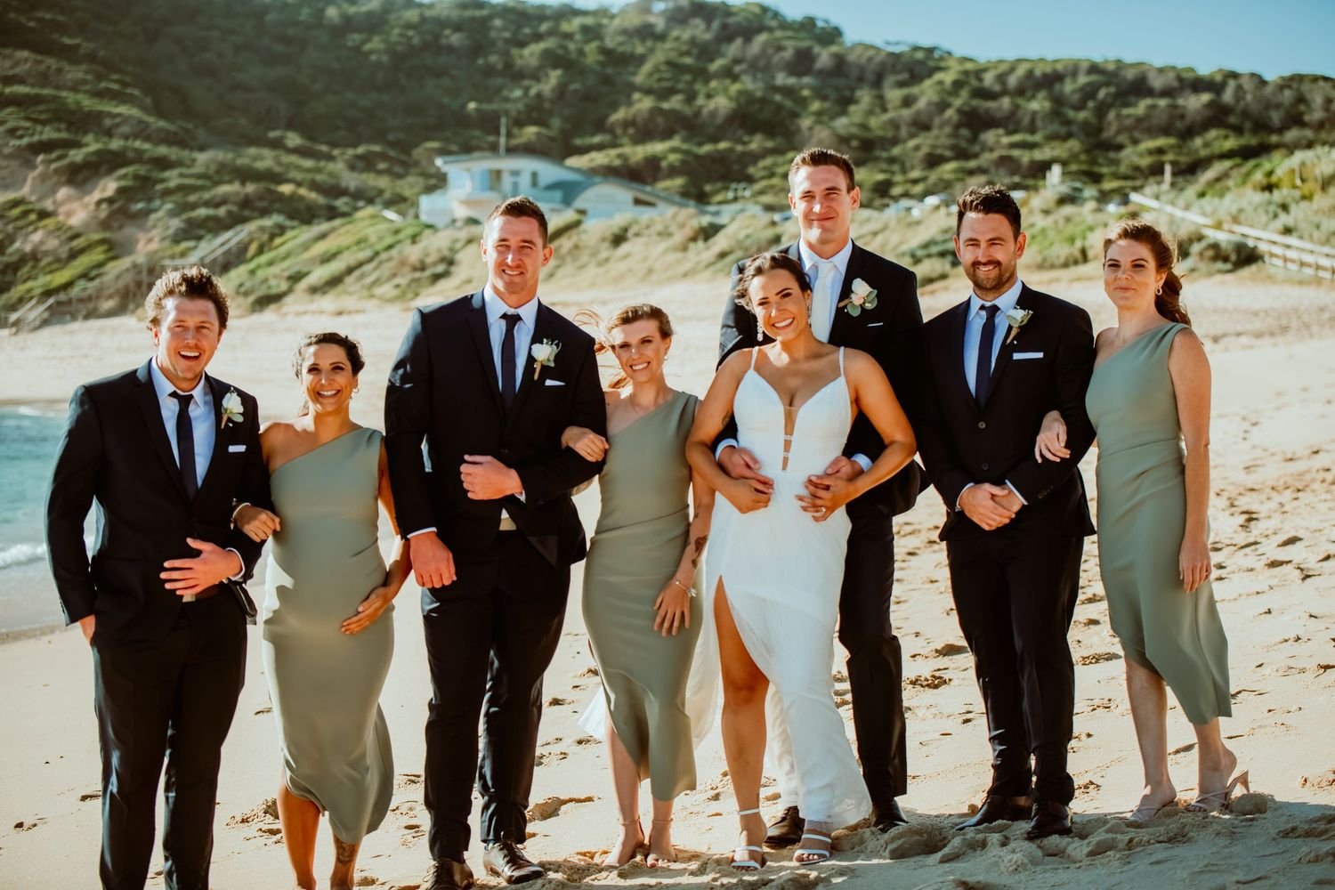 Wedding party in formal attire poses together on a scenic beach with hills in the background on a sunny day.