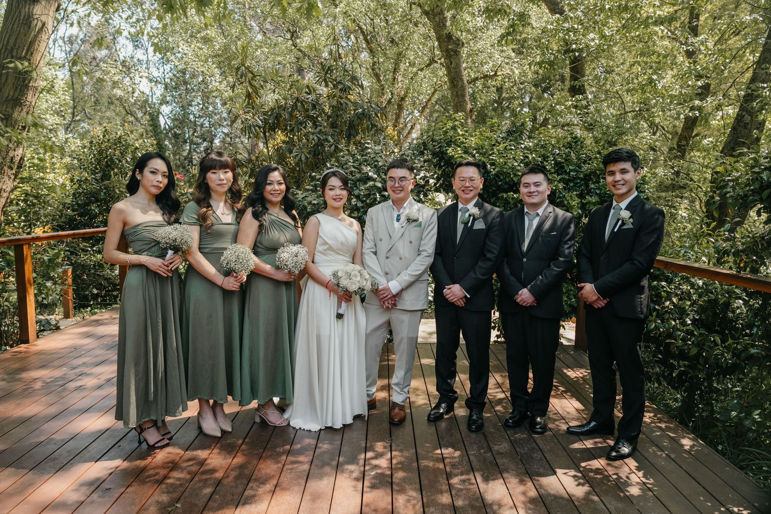 Wedding party poses outdoors on wooden deck surrounded by lush greenery on a sunny day.