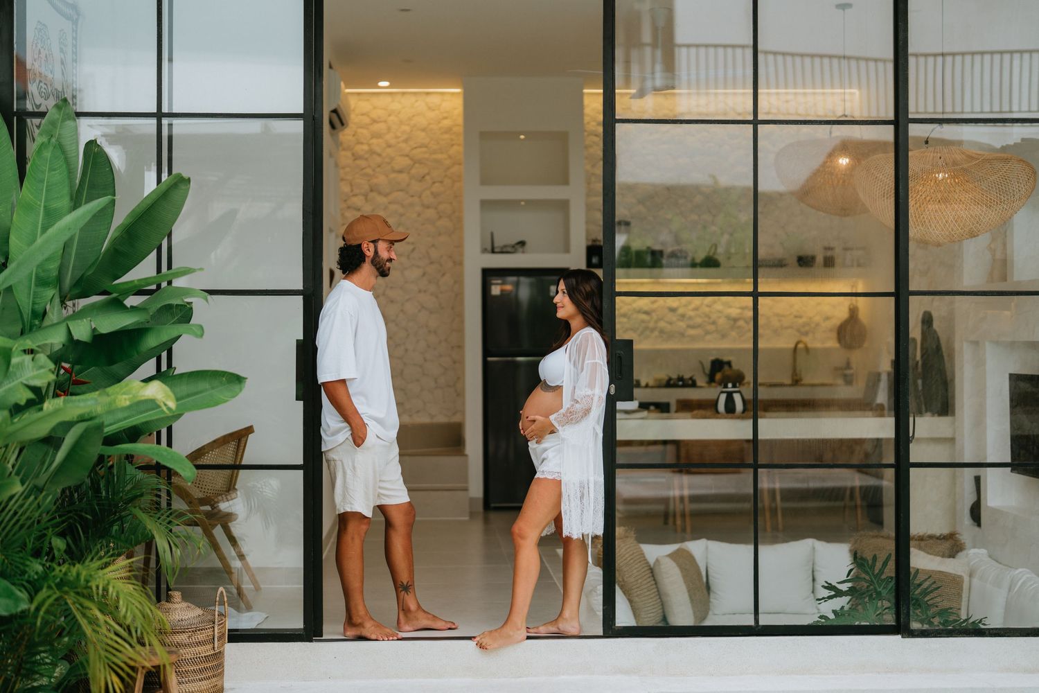 Two people in white outfits stand in conversation at a modern glass-walled home entrance.