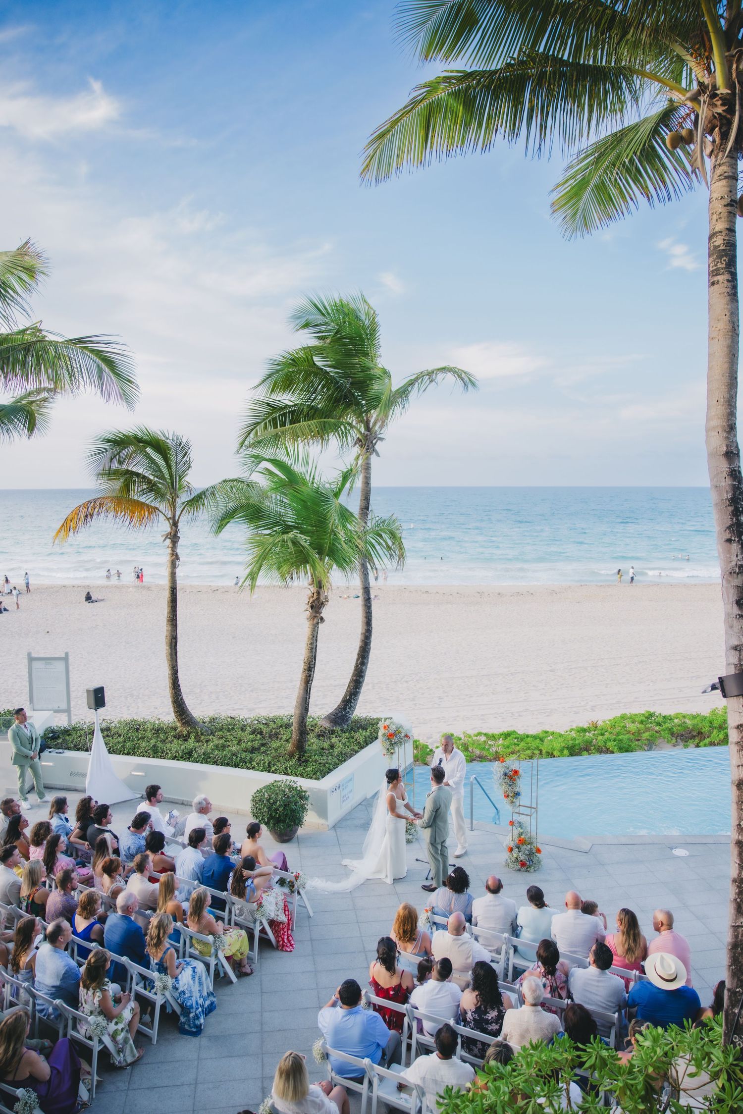 Wedding guests gather on an oceanfront terrace with palm trees and beach views during a tropical seaside ceremony.