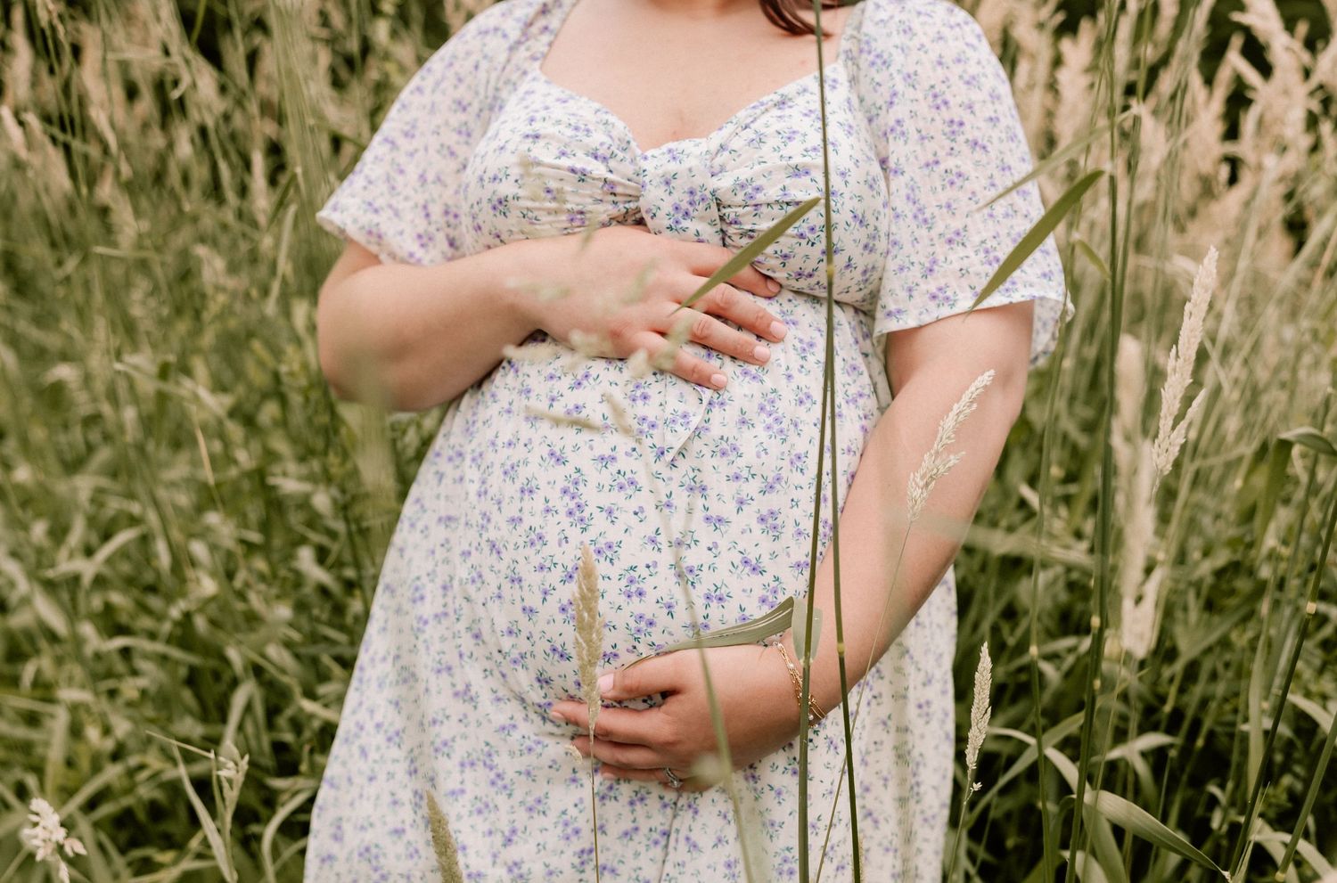 A person in a floral dress gently touches tall wild grass and plants in a sunny meadow field outdoors.