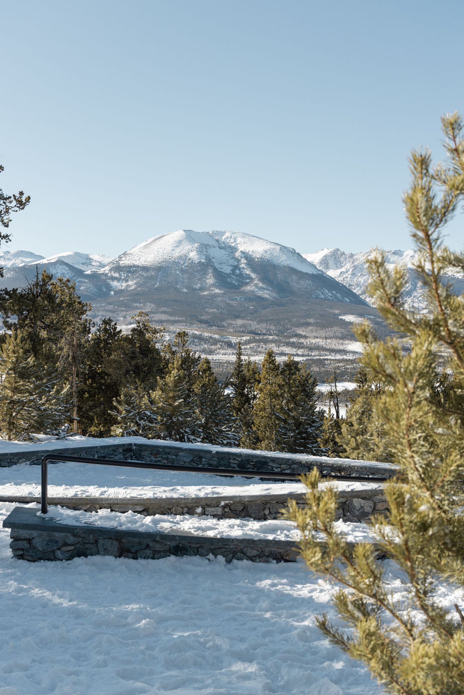 Snow-covered mountain peaks rise against a clear blue sky with evergreen trees dotting the winter landscape.