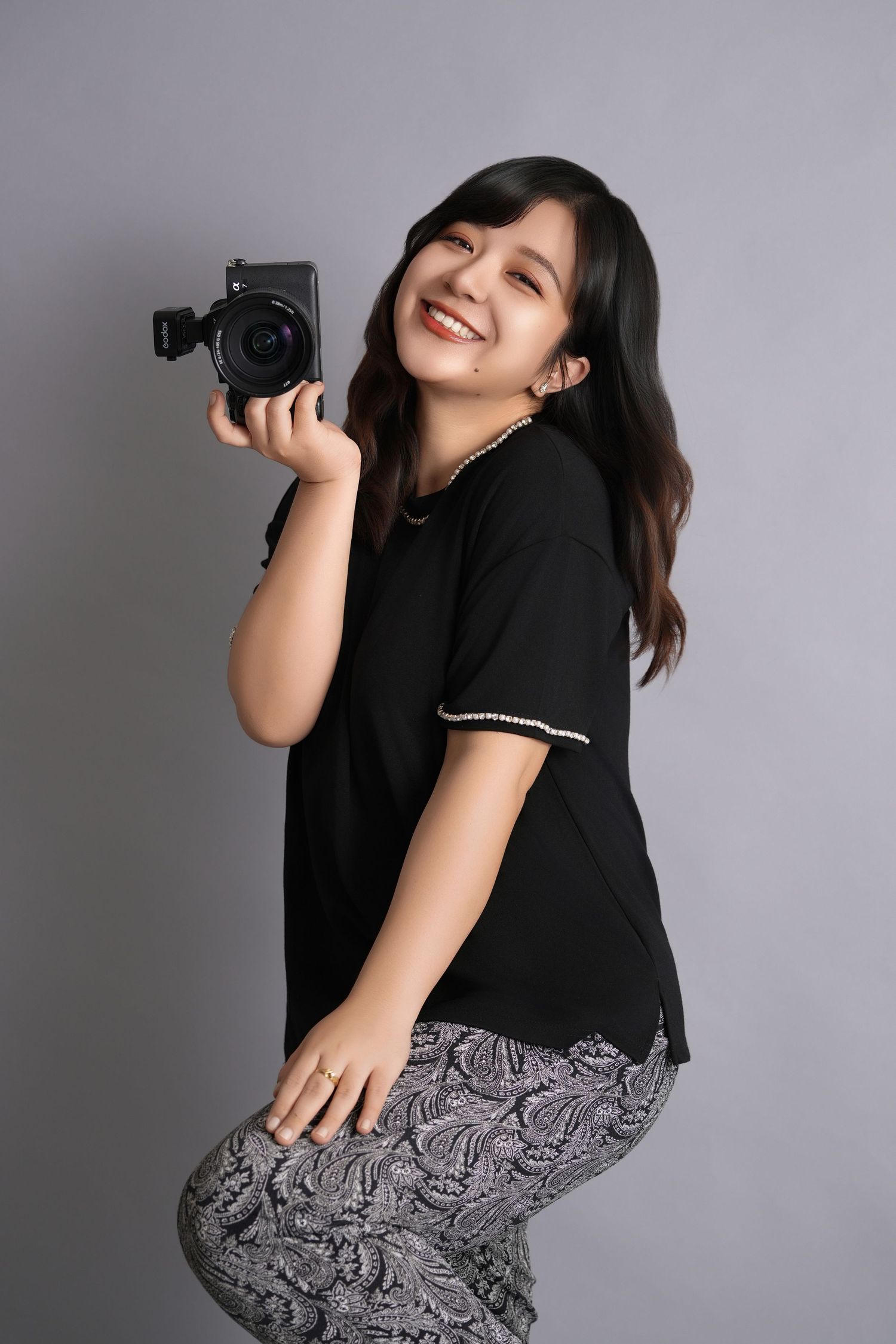 Woman in black top and patterned skirt poses playfully against grey background with camera.