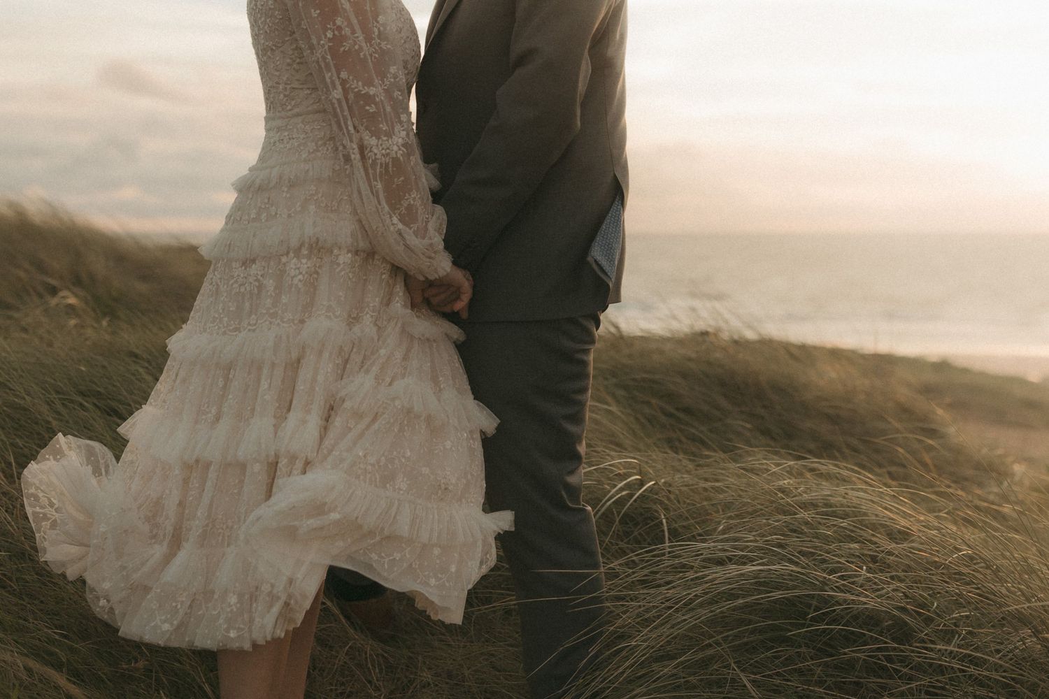 A couple in wedding attire walks through windswept coastal grass at sunset near rugged cliffs.